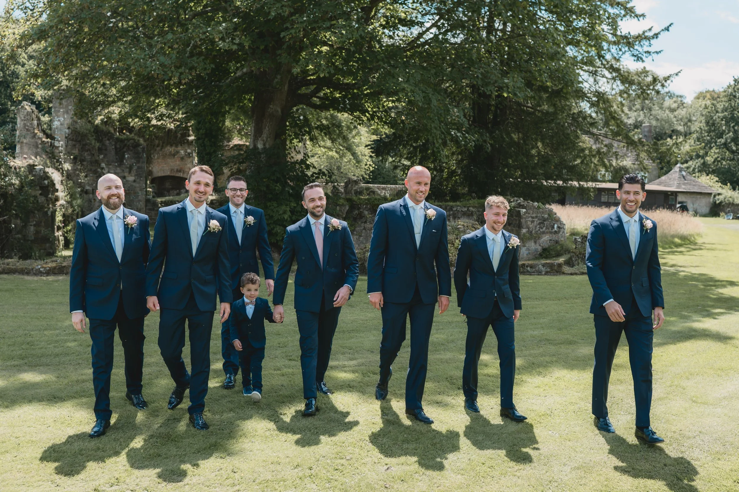 Groom and groomsmen in navy suits walking on a lawn during a wedding celebration outdoors.