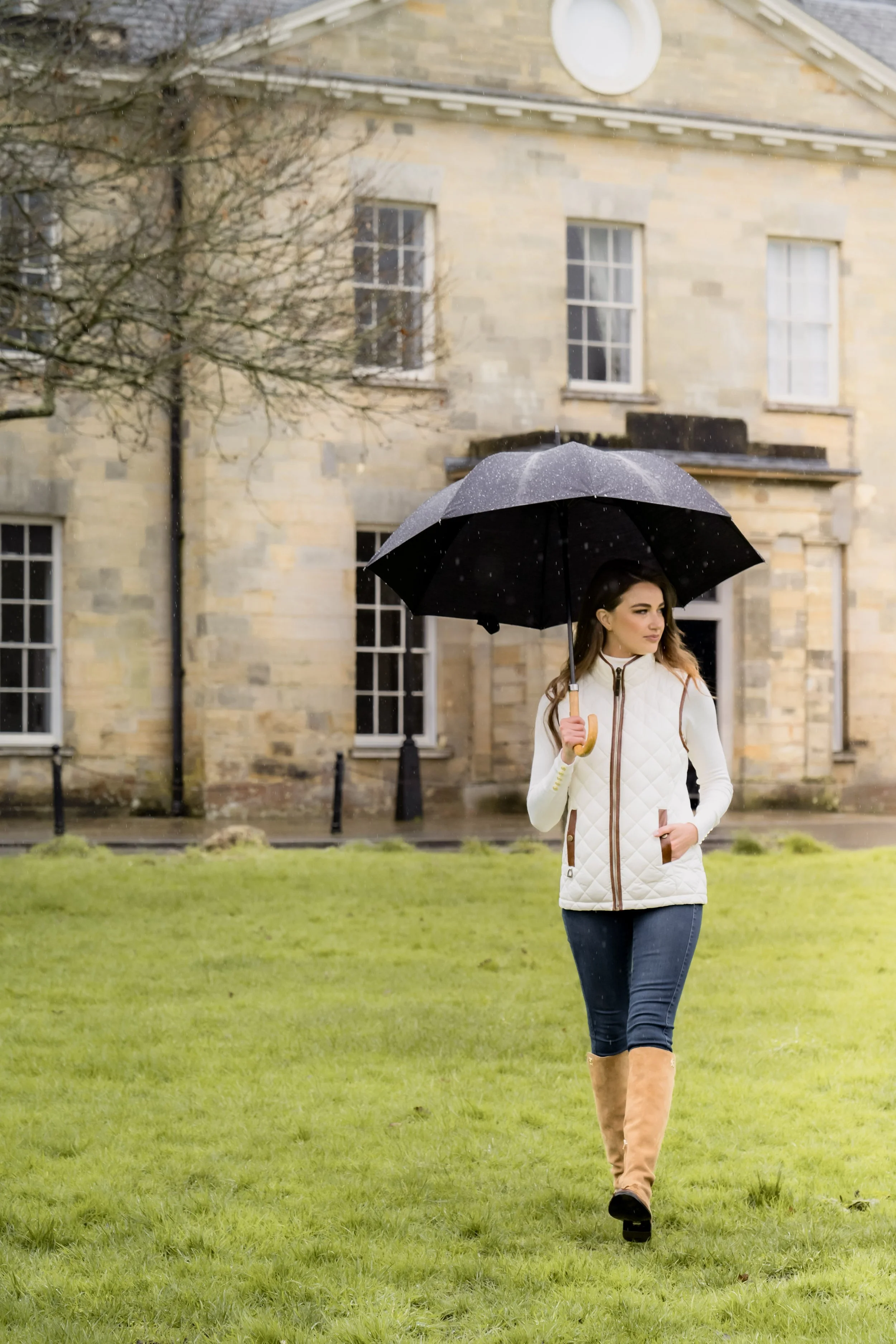Woman walking on a grassy lawn holding a black umbrella during rain with a stone building in the background.