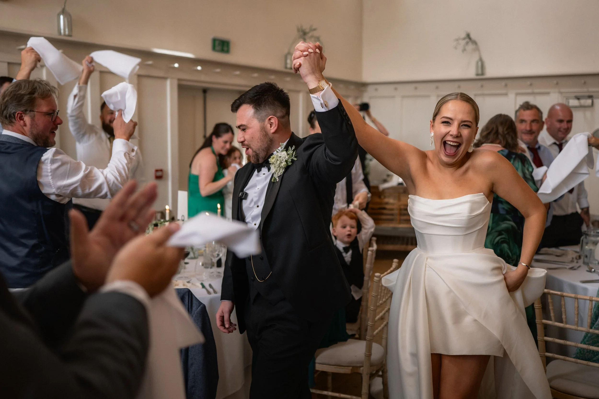 A bride and groom celebrate at a wedding reception, holding hands and smiling with joy. The bride is wearing a strapless white dress, and the groom is in a black tuxedo. Guests are dancing and cheering in the background.