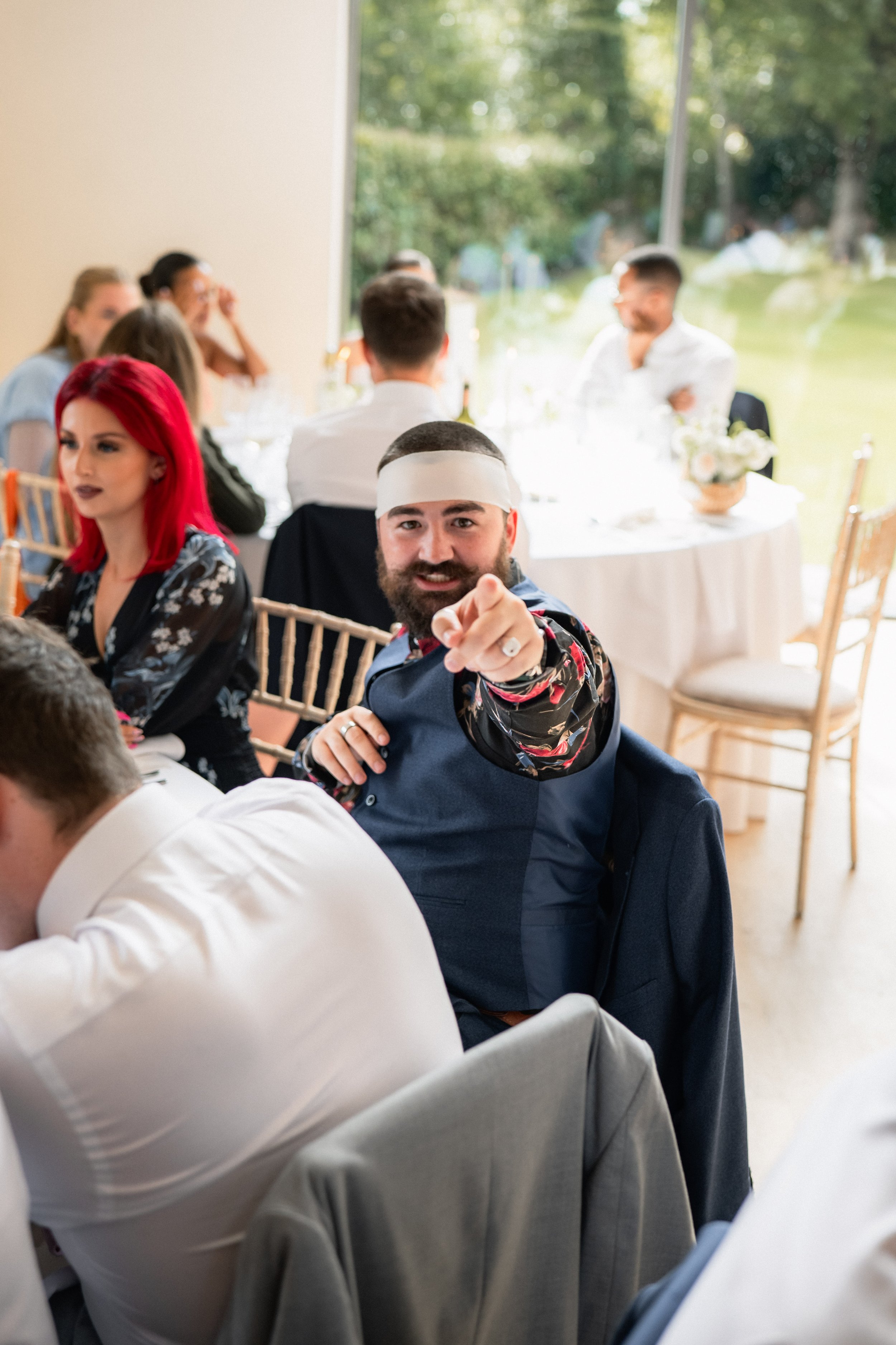 Man with a beard, wearing a paper crown and a floral shirt, pointing towards the camera at a formal event, with a woman with bright red hair and a black dress seated nearby. In the background, other guests are seated at tables in a decorated indoor s