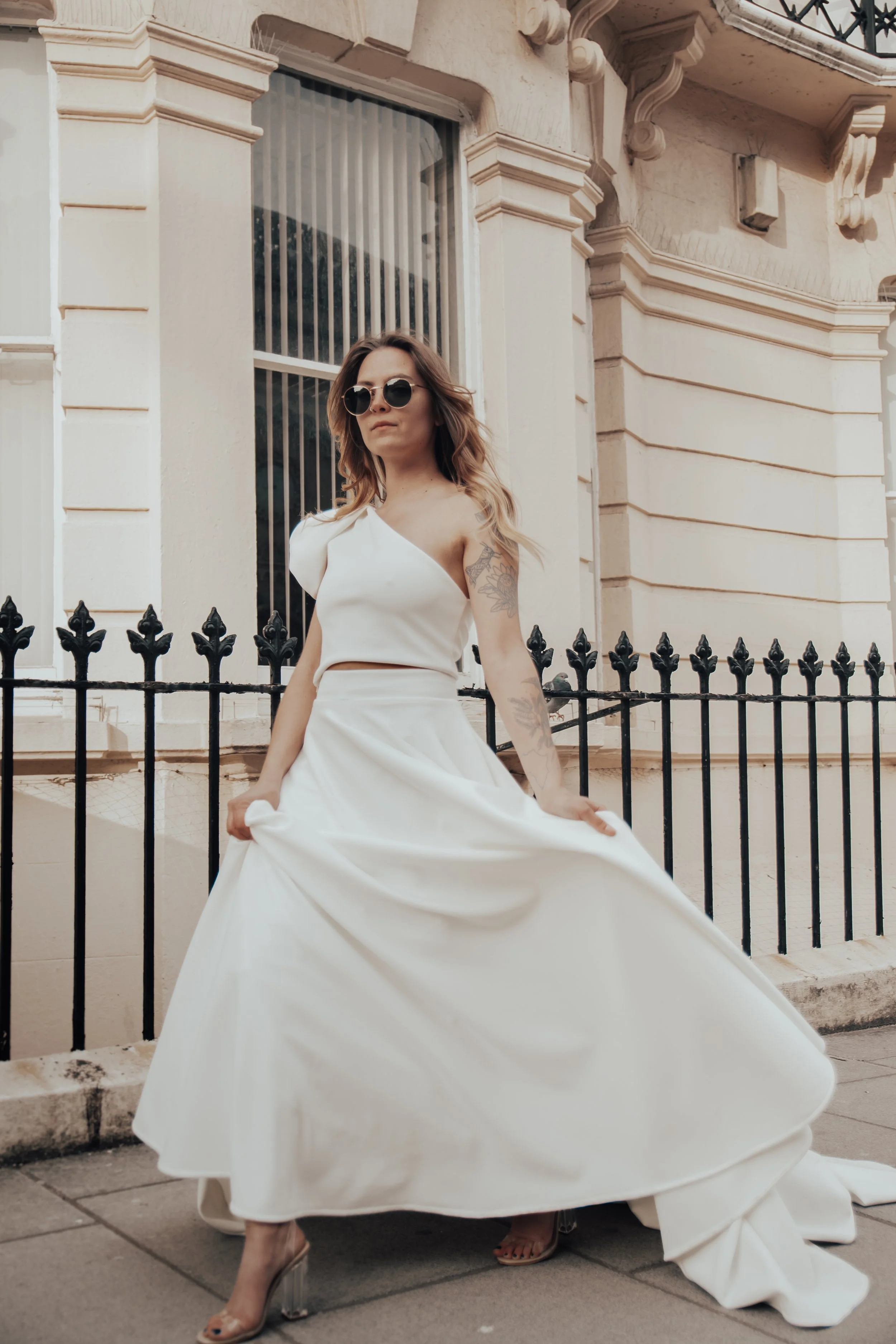 A woman in a white wedding dress with sunglasses standing in front of a building with tall windows and a black iron fence.