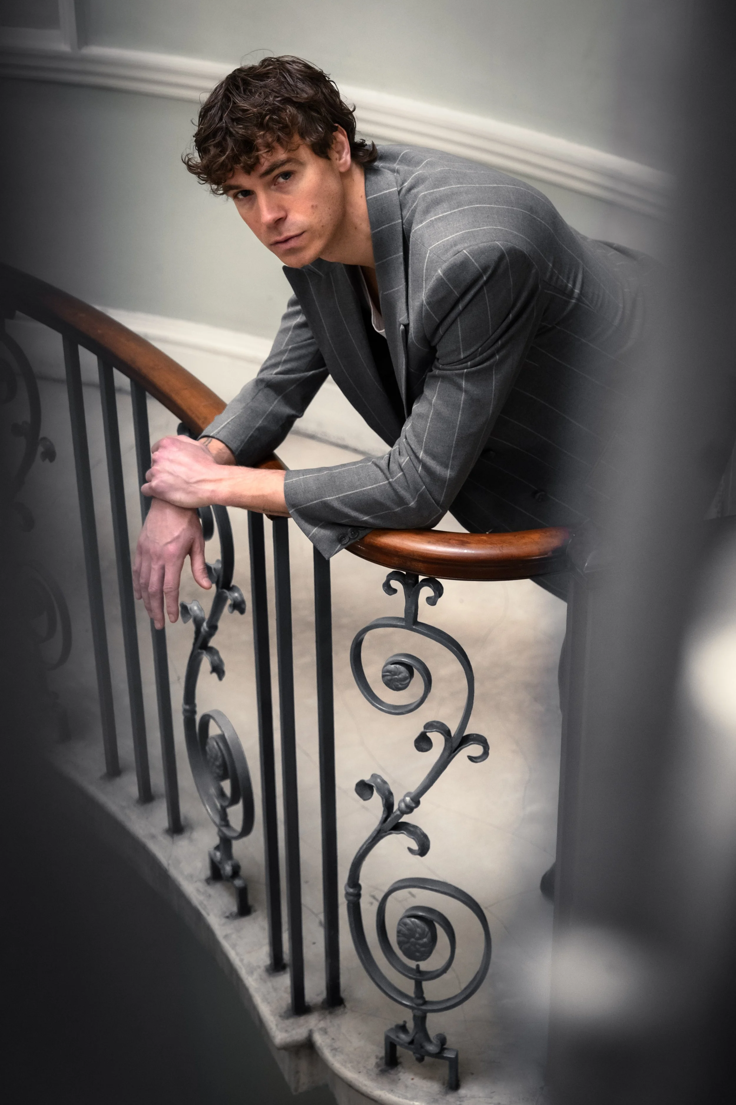 A young man with curly hair leaning on a staircase railing, looking at the camera.