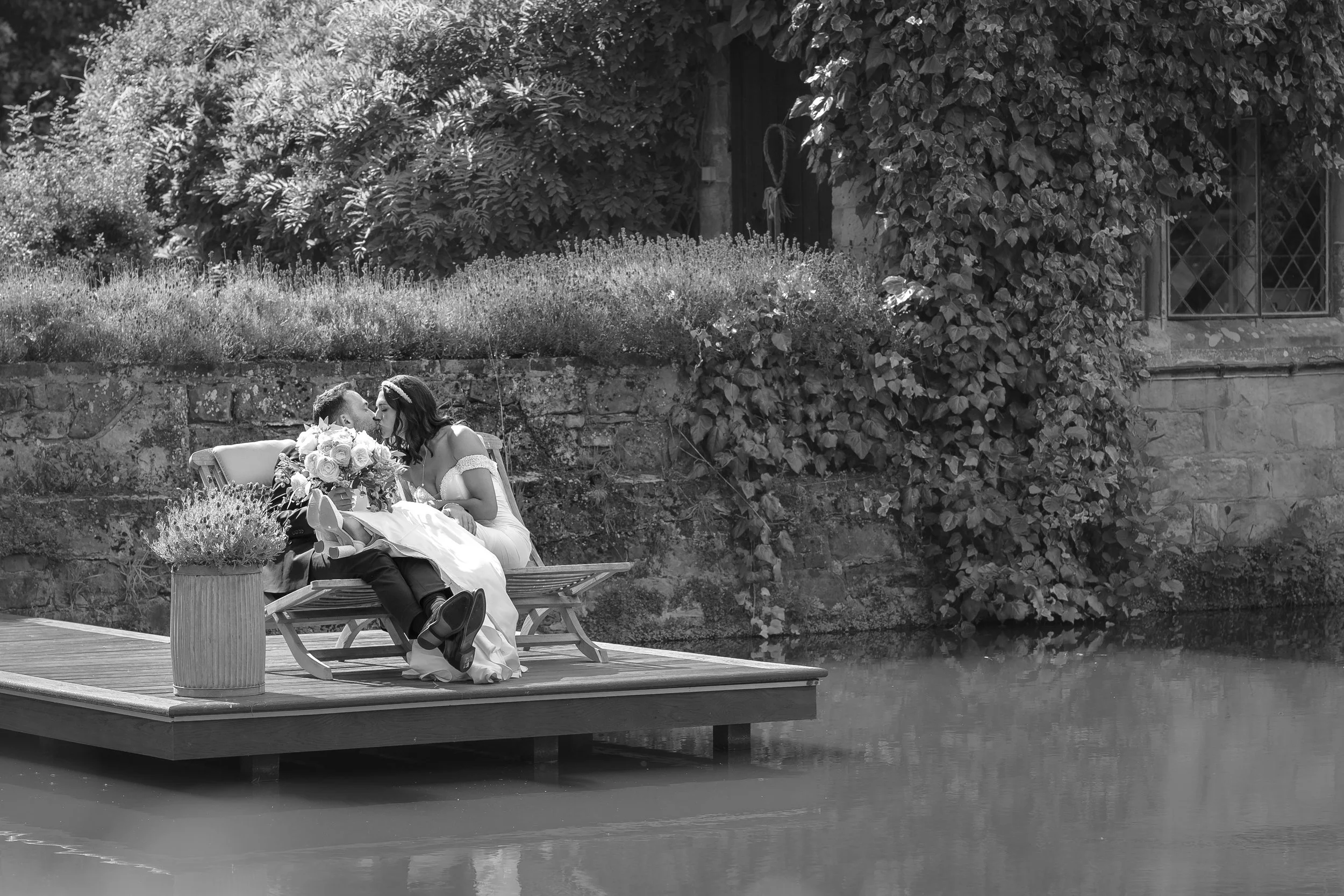 Black and white photo of a woman and child sitting on a bench on a dock by the water, sharing a kiss. The woman is in a white dress and holding a bouquet of flowers, while the child is dressed in dark clothing. There are plants and a stone wall in th