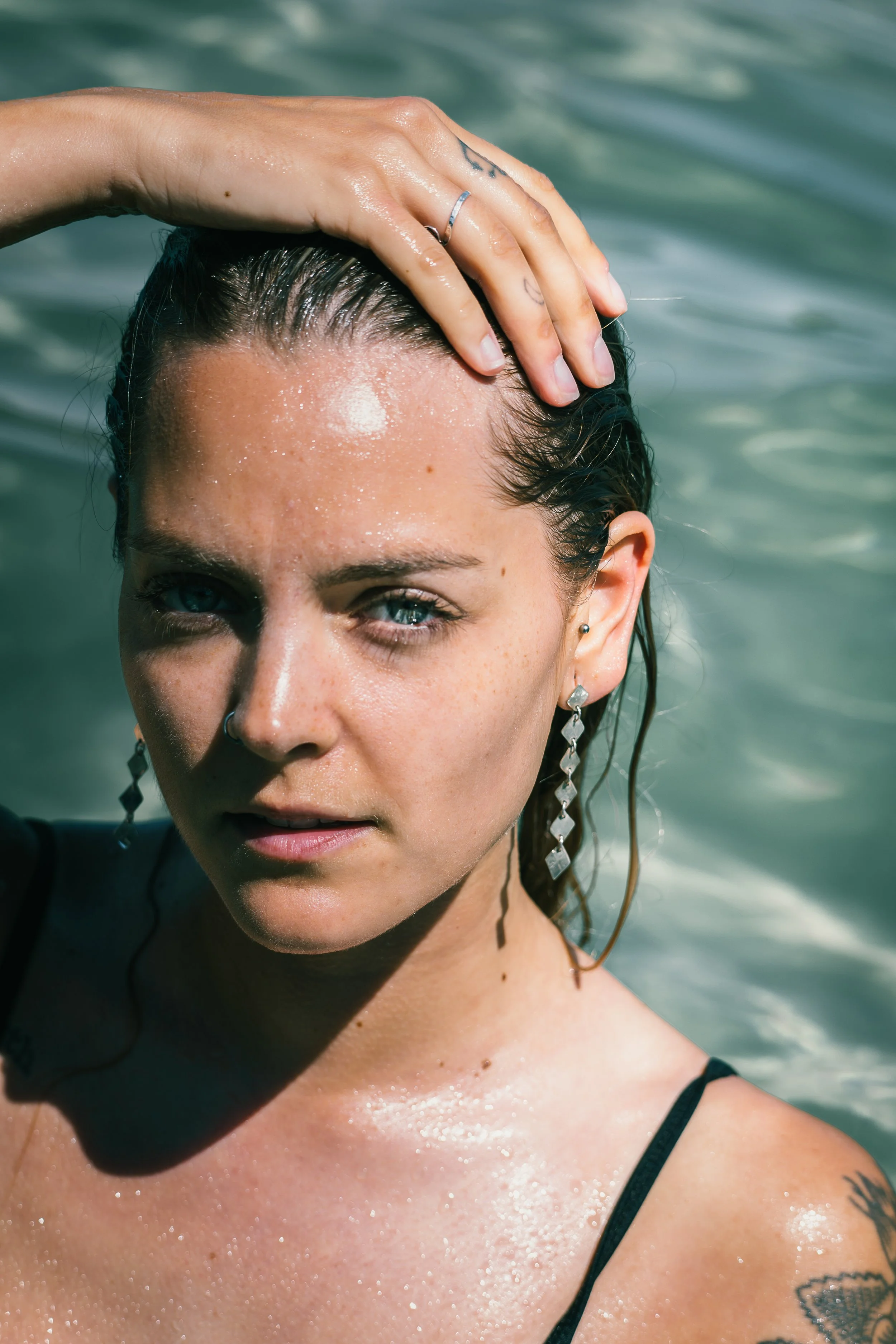 A woman with wet hair and blue eyes posing in front of water, wearing earrings and a black strap top, with her hand on her head.
