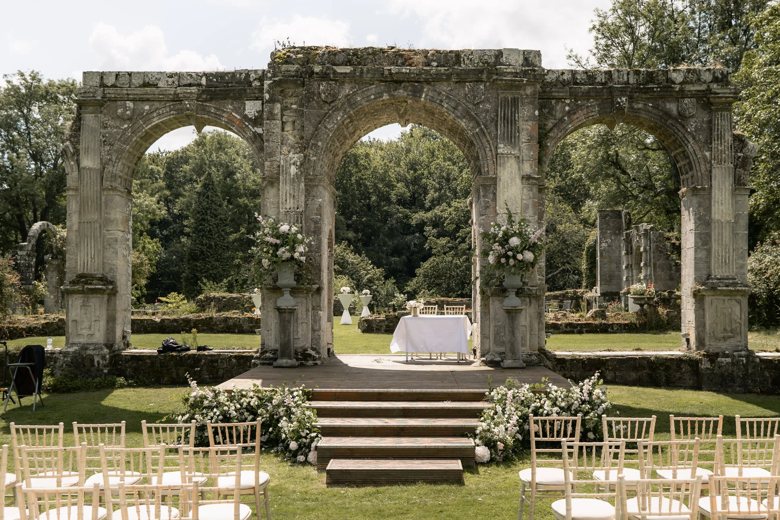 Outdoor wedding ceremony setup with white chairs, floral arrangements, table, and stone ruins with arches on a grassy area.