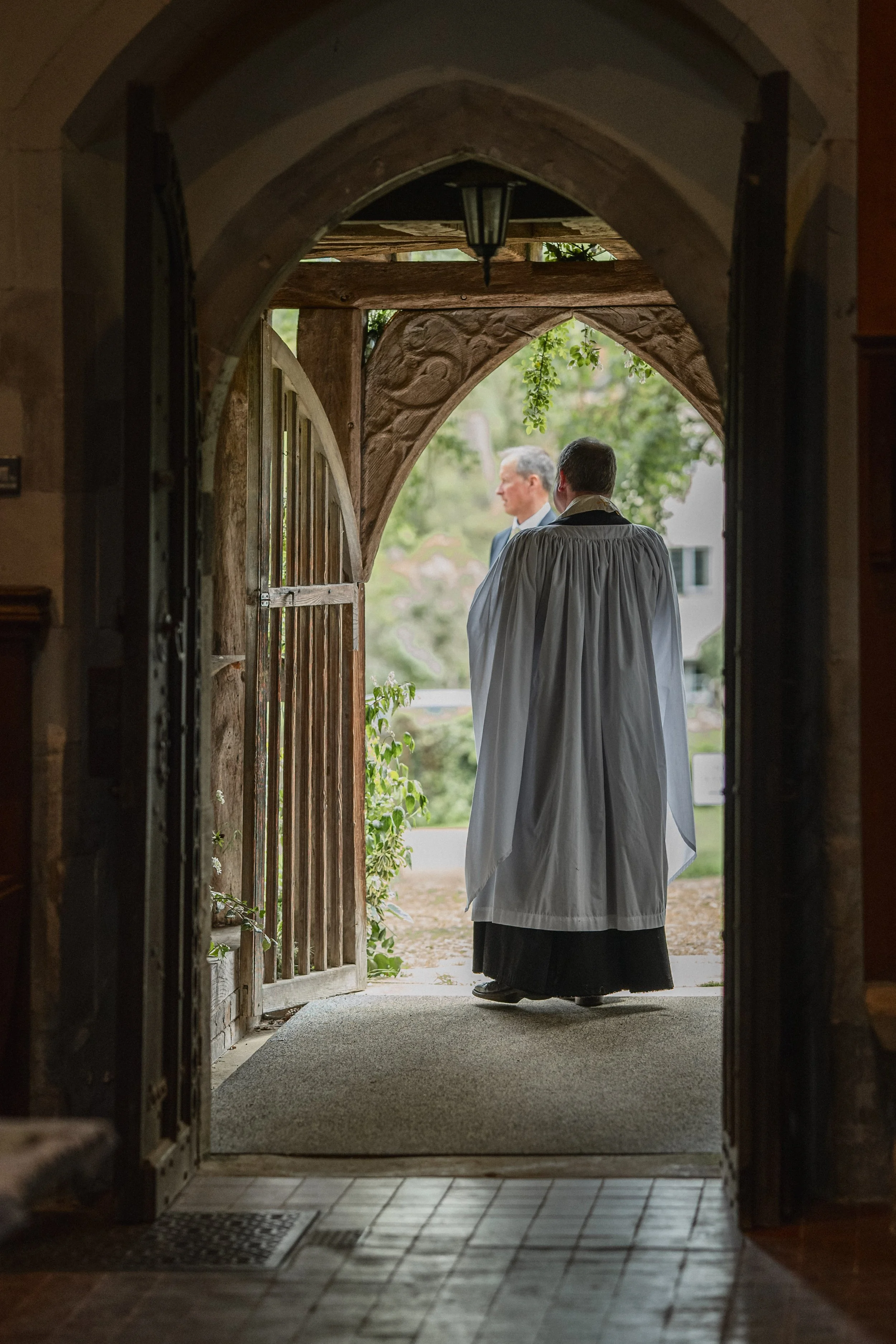 A person dressed in a religious robe standing at an open wooden gate outside, with a man in a suit visible in the background among green trees.