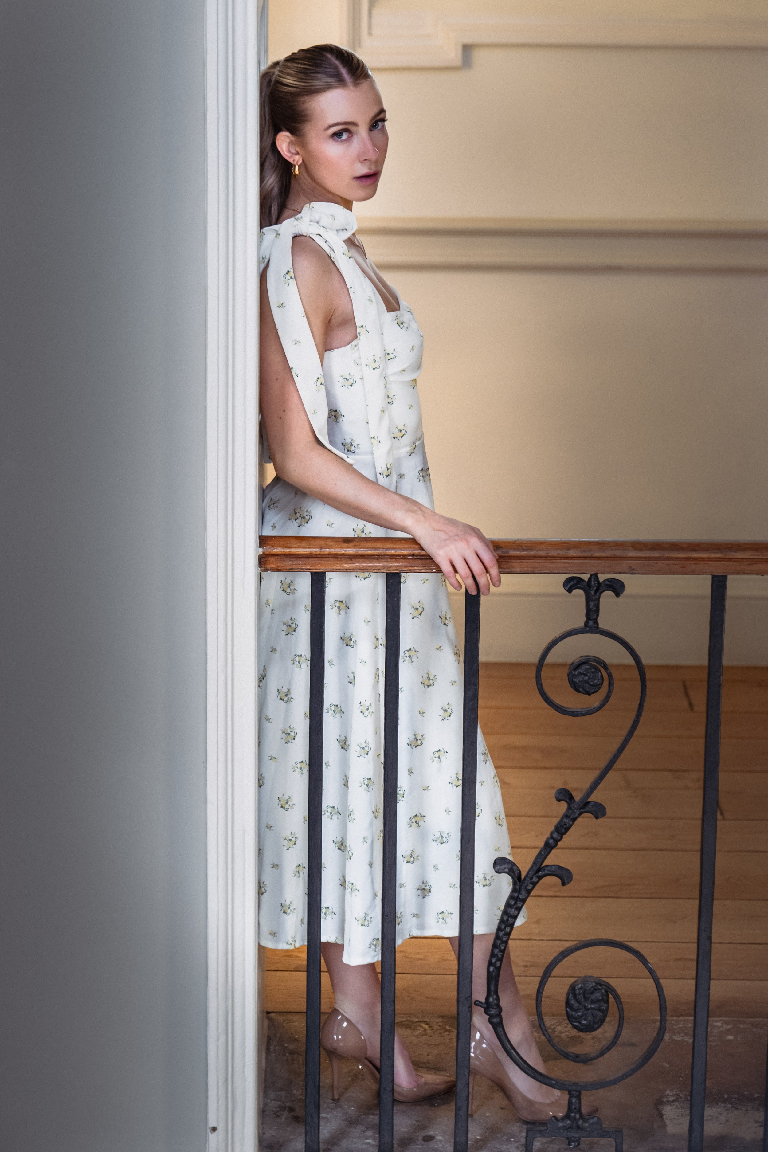 Young woman with brown hair in a ponytail, wearing a white floral dress with tie-up shoulders, standing behind a metal railing with a wooden handrail, inside a room with cream-colored walls and wooden floors.