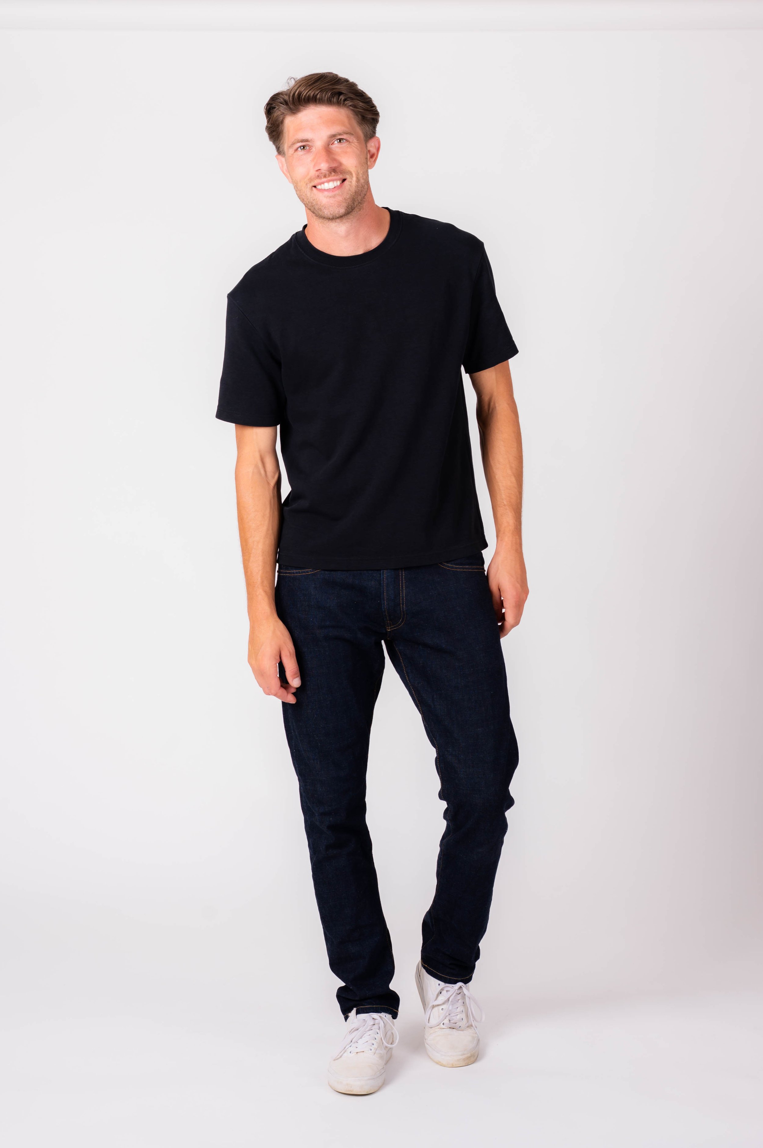 A smiling man with light brown hair, wearing a black T-shirt, dark jeans, and white sneakers, standing against a plain white background.