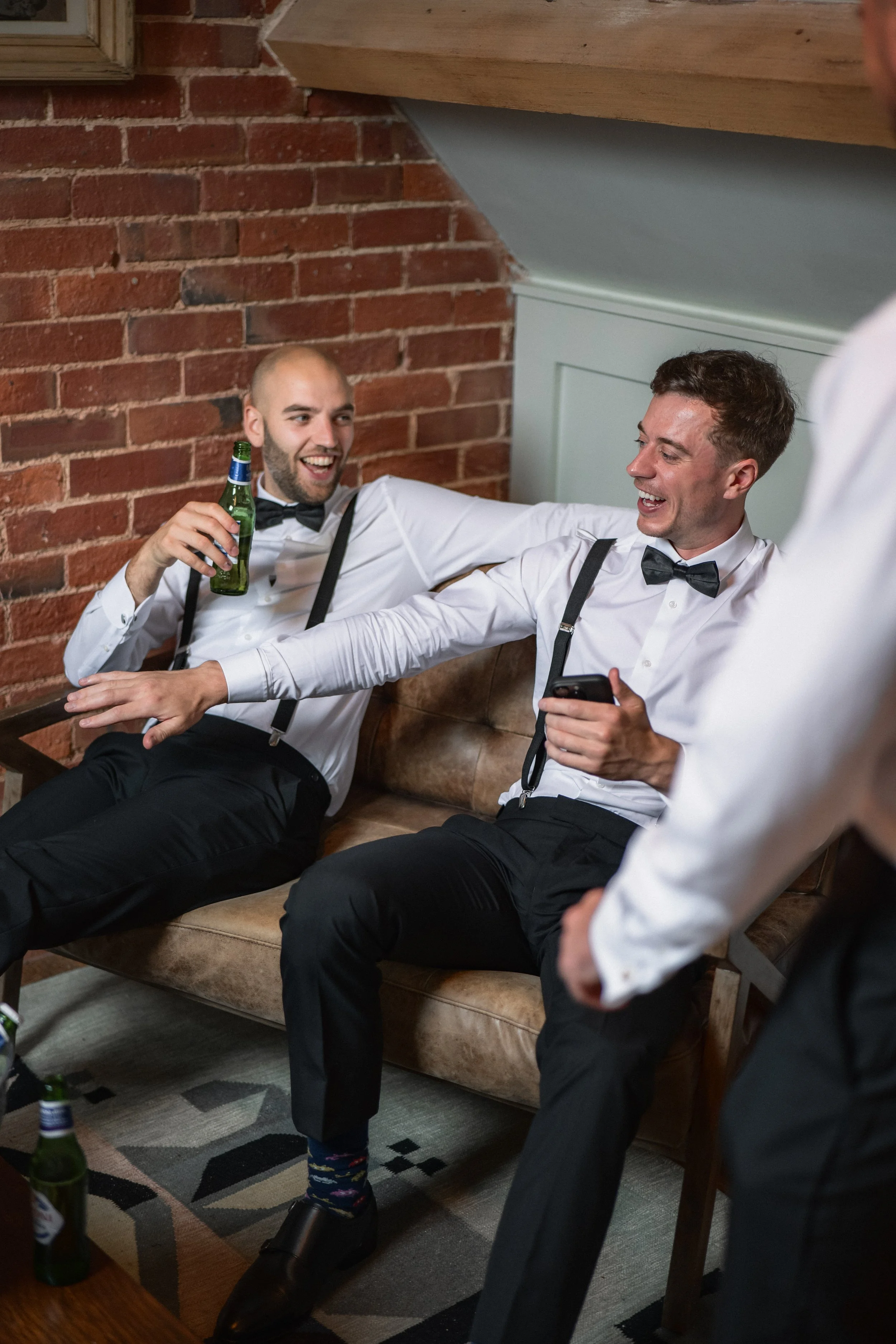Two men in formal attire, white shirts, and black bow ties, sitting on a brown leather sofa and laughing. One man holds a beer bottle, and the other man is holding a smartphone. A third person is partially visible, standing in front of them. The sett
