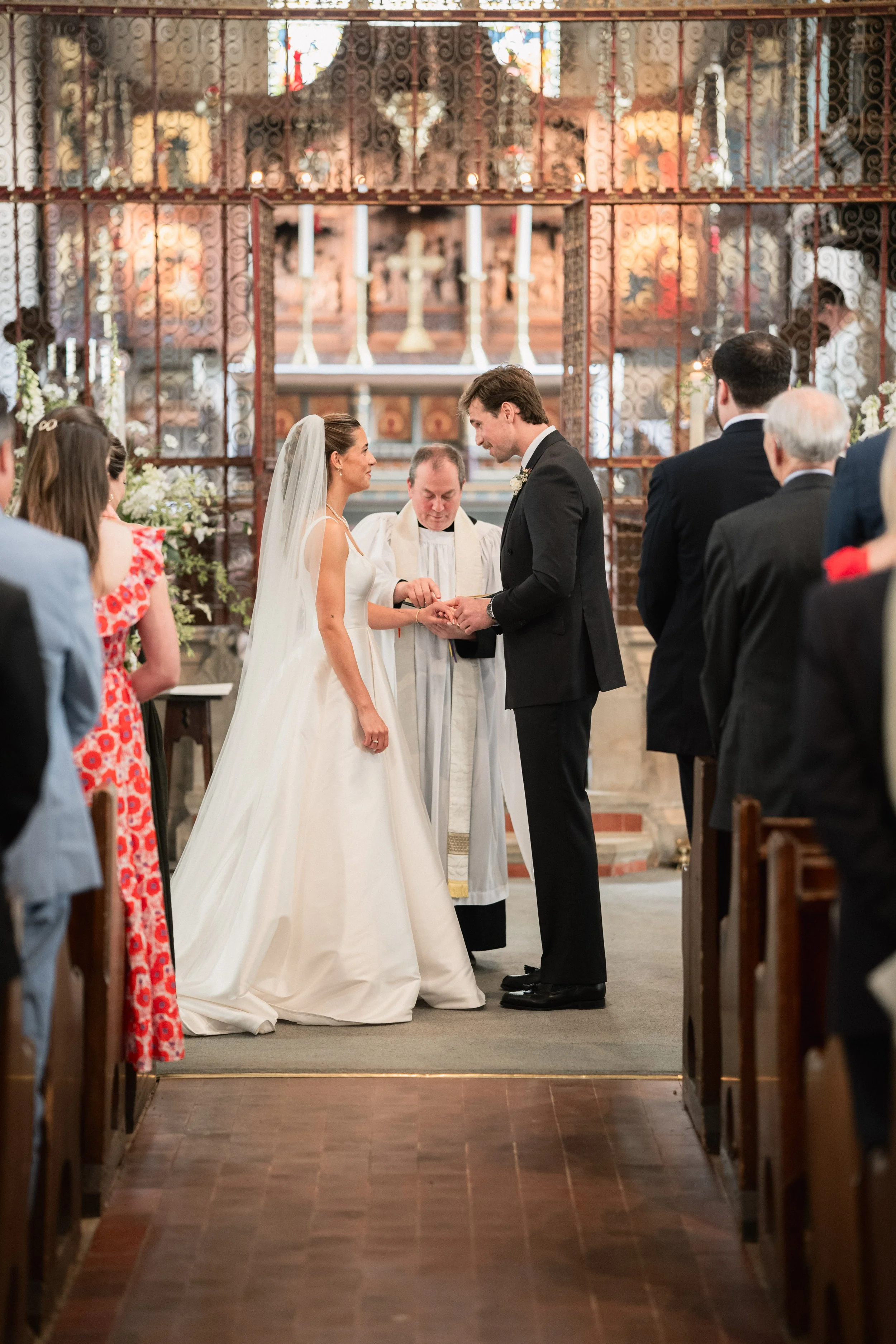 A wedding ceremony inside a church with the bride and groom exchanging vows, surrounded by officiant and guests, with an ornate altar in the background.