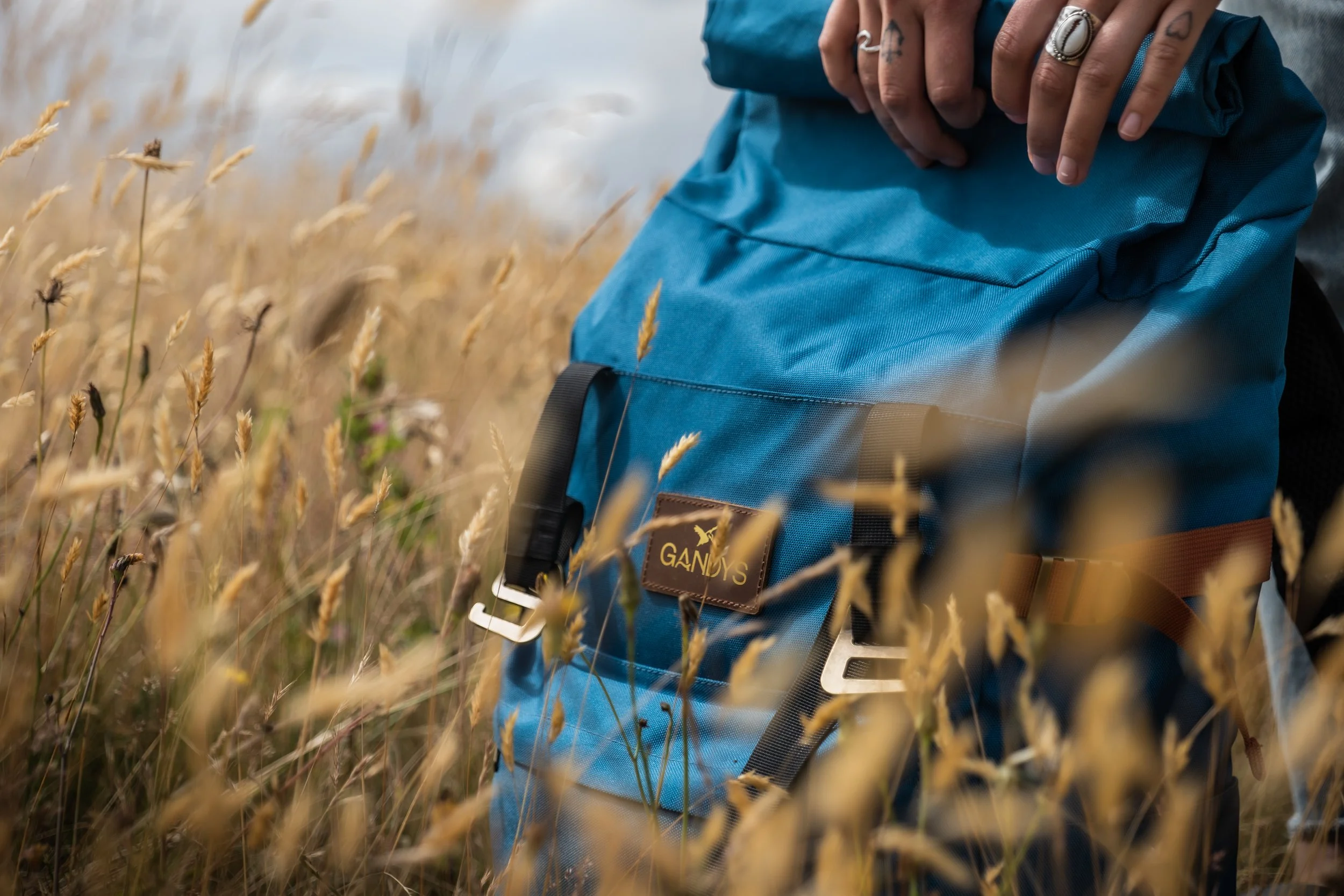 Close-up of a blue canvas backpack with a leather patch labeled "GANGES" amidst tall, dry grass, with a person's hand wearing rings resting on top of the backpack.