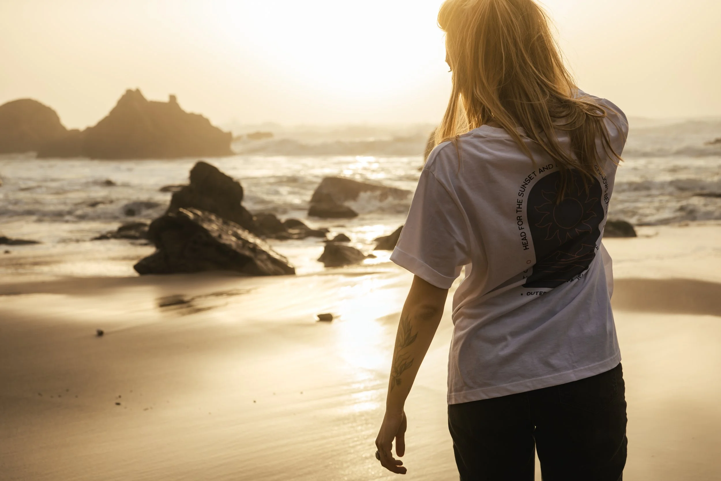 A person with long hair standing on a beach at sunset, facing the ocean with rocks in the water and sun in the background.