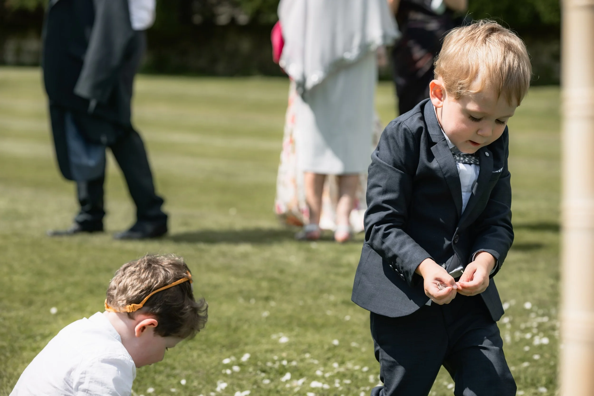 Two young boys in formal attire on a grassy lawn, with adults in the background at an outdoor event.