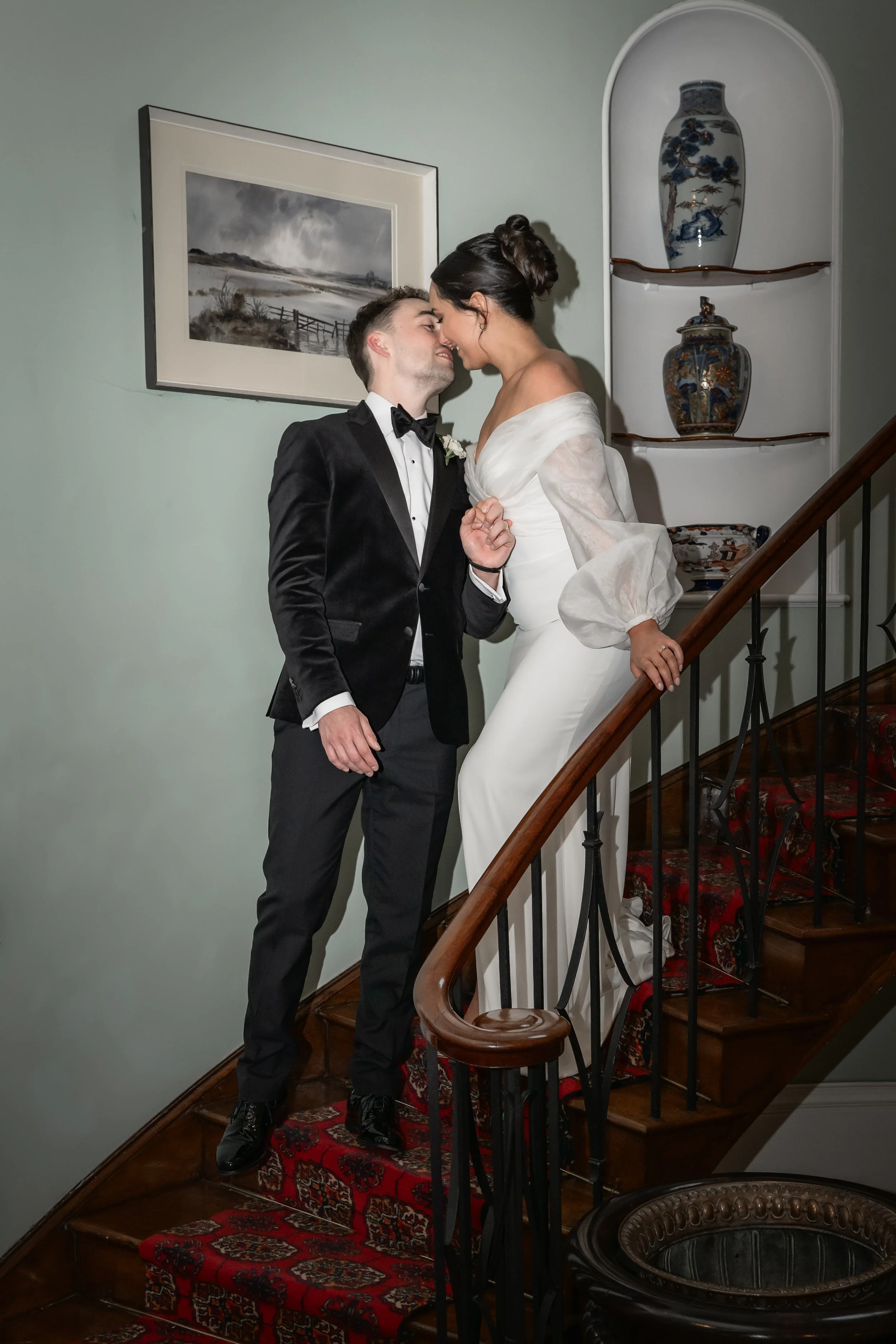 A bride and groom share a moment on a staircase during their wedding. The groom wears a black tuxedo, and the bride wears a white wedding dress. They are leaning in close, touching foreheads.