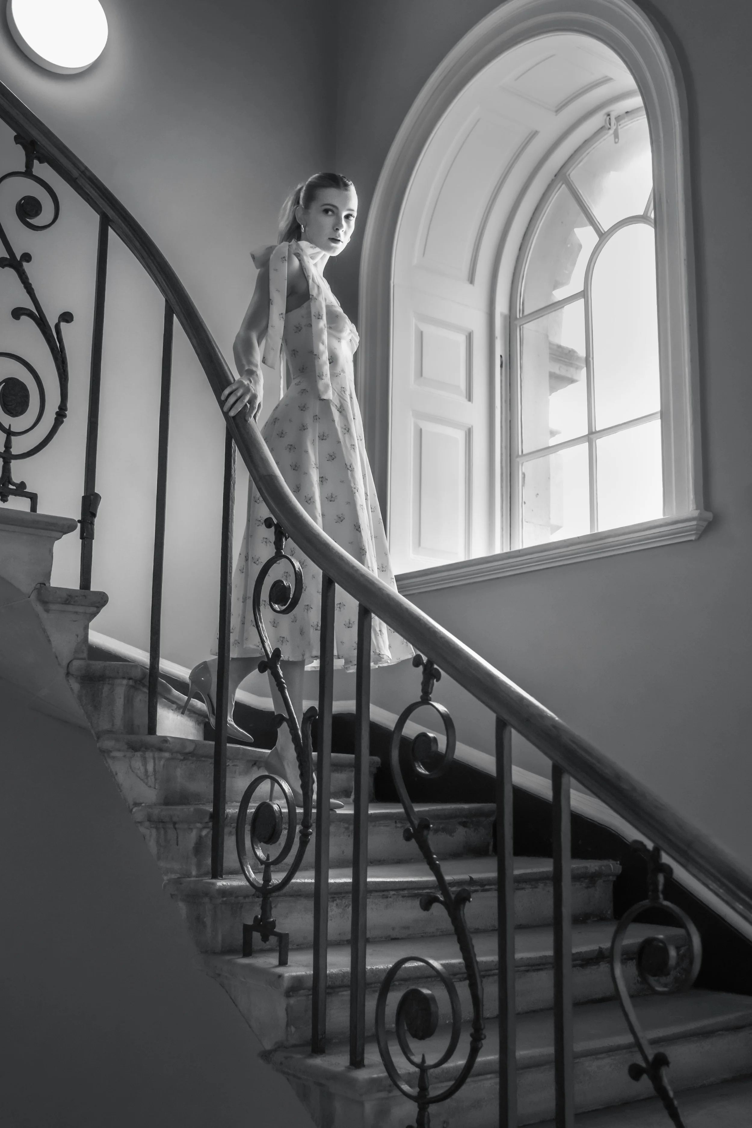 A woman in a vintage floral dress standing on a staircase near a large arched window, with sunlight streaming in, in a black and white photo.
