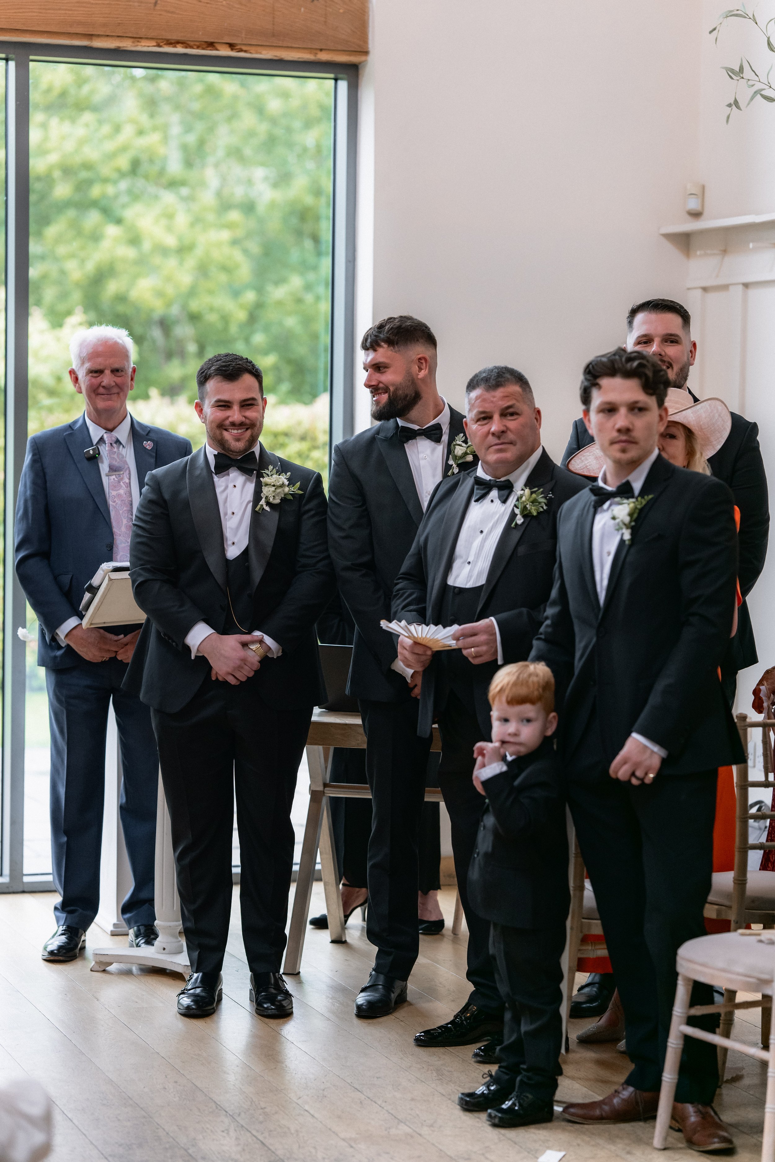 Group of men dressed in black tuxedos and bow ties, standing indoors during a wedding ceremony. One young boy in a suit is in the front, and a woman with a large hat is partially visible behind them. The background shows a large window with trees out