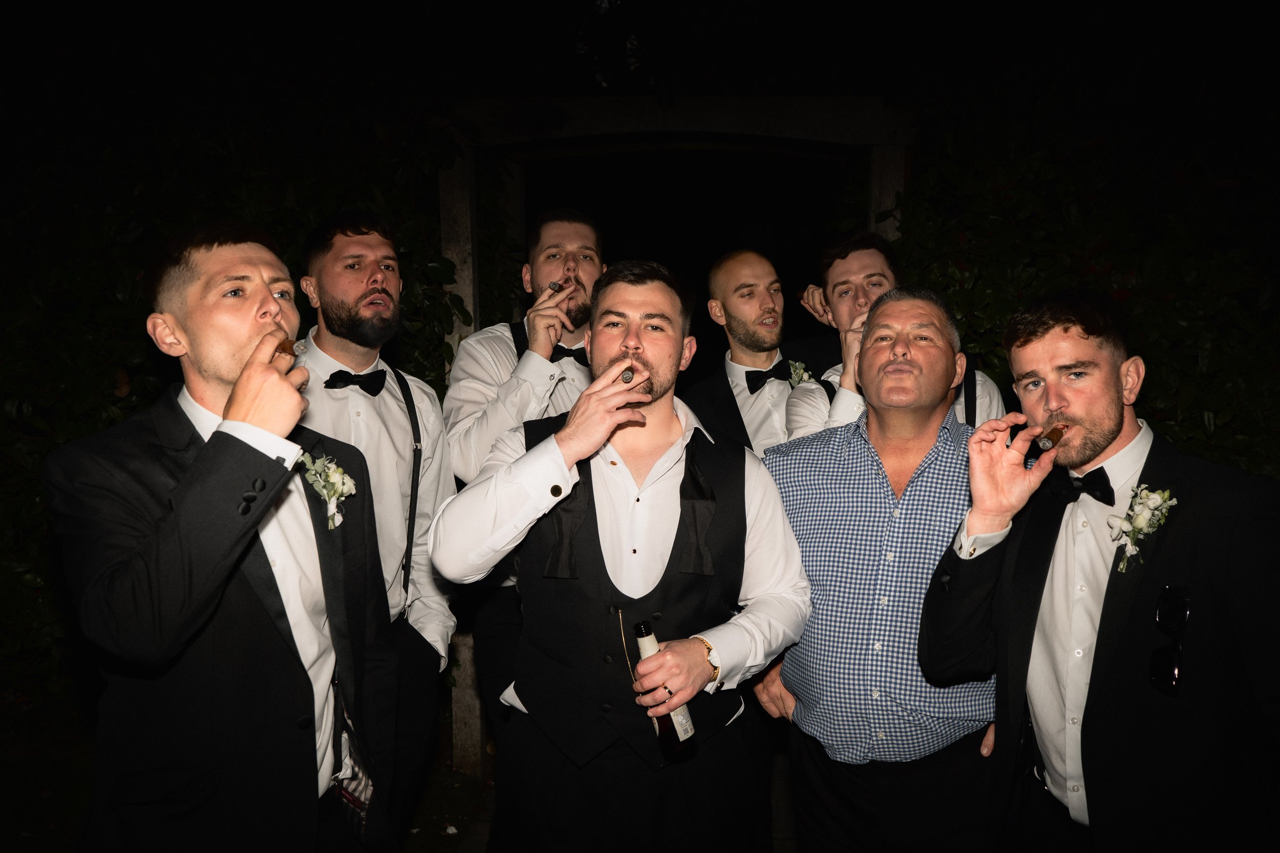 A group of men in tuxedos and dress shirts, some with suspenders, smoking cigars at night.