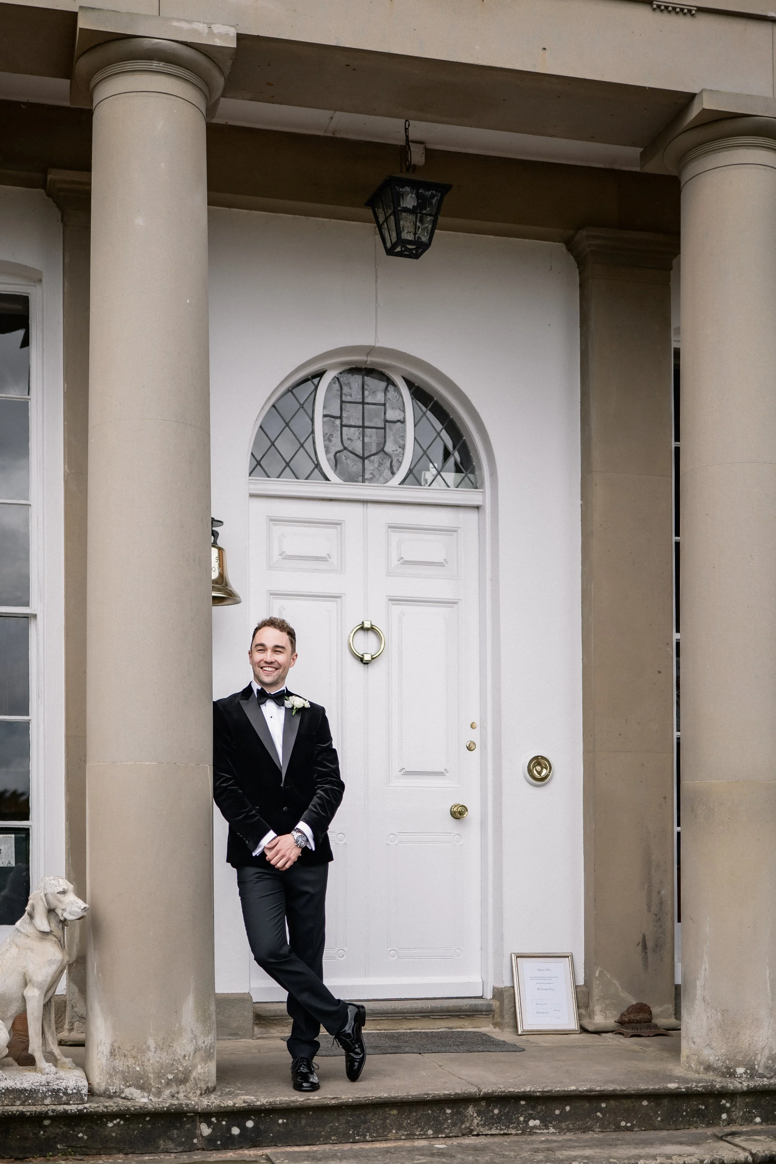 A man in a black tuxedo with a bow tie and boutonniere standing on the front porch of a house, smiling, with one leg crossed over the other, in front of a white door with a decorative window and doorbell.