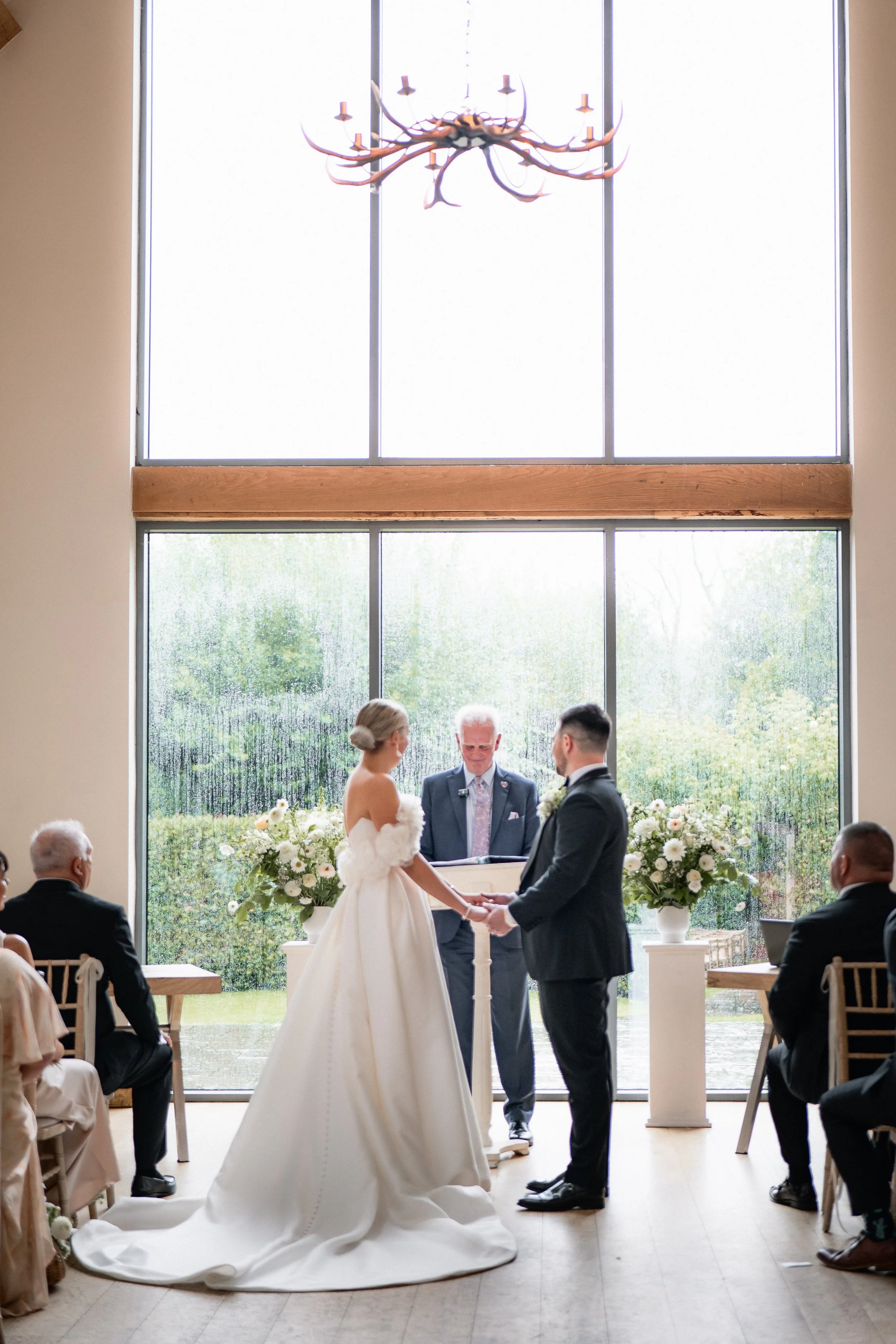 A wedding ceremony with a bride and groom holding hands, standing in front of an officiant in a bright, indoor space with large glass windows, floral arrangements, and seated guests.