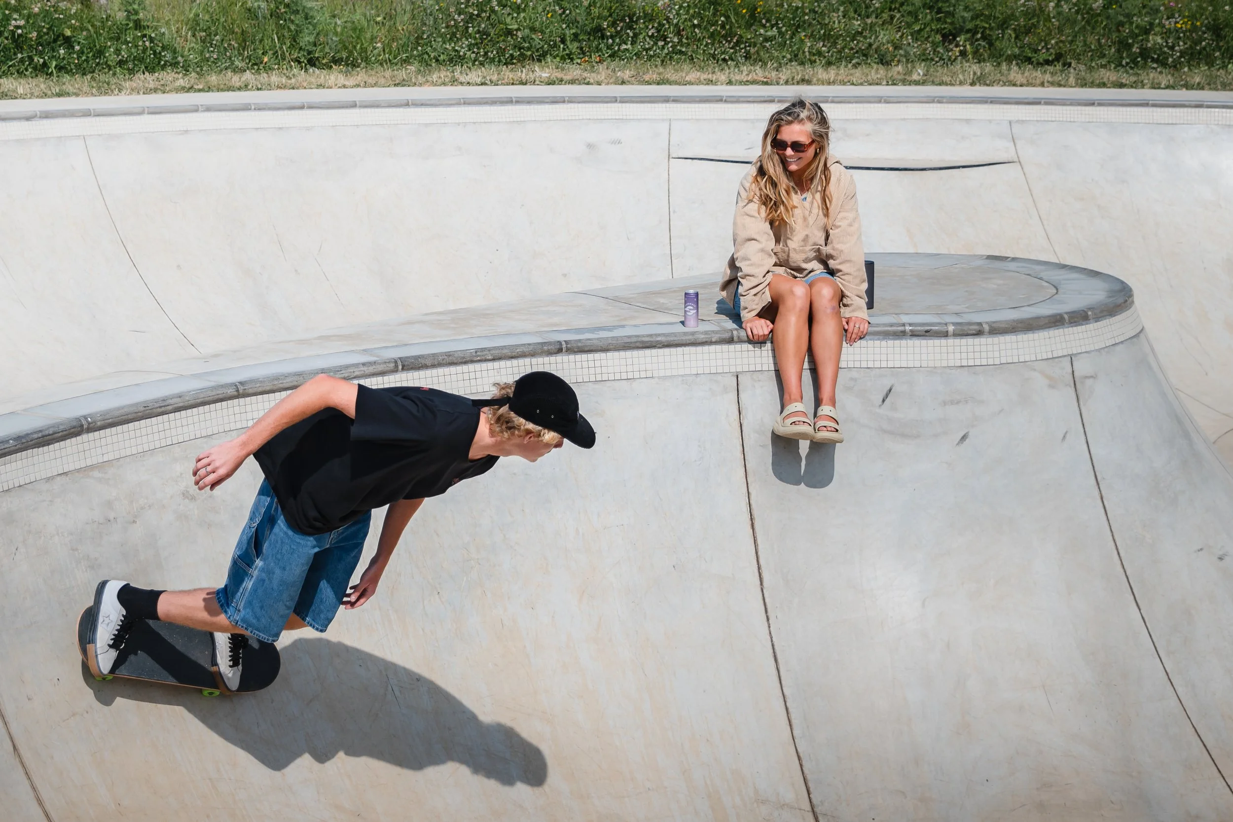 A young man skateboarding in a concrete skate bowl with a girl sitting on the edge, watching him, smiling. The girl has blonde hair, sunglasses, and is wearing a beige jacket and sandals. A purple drink is next to her.