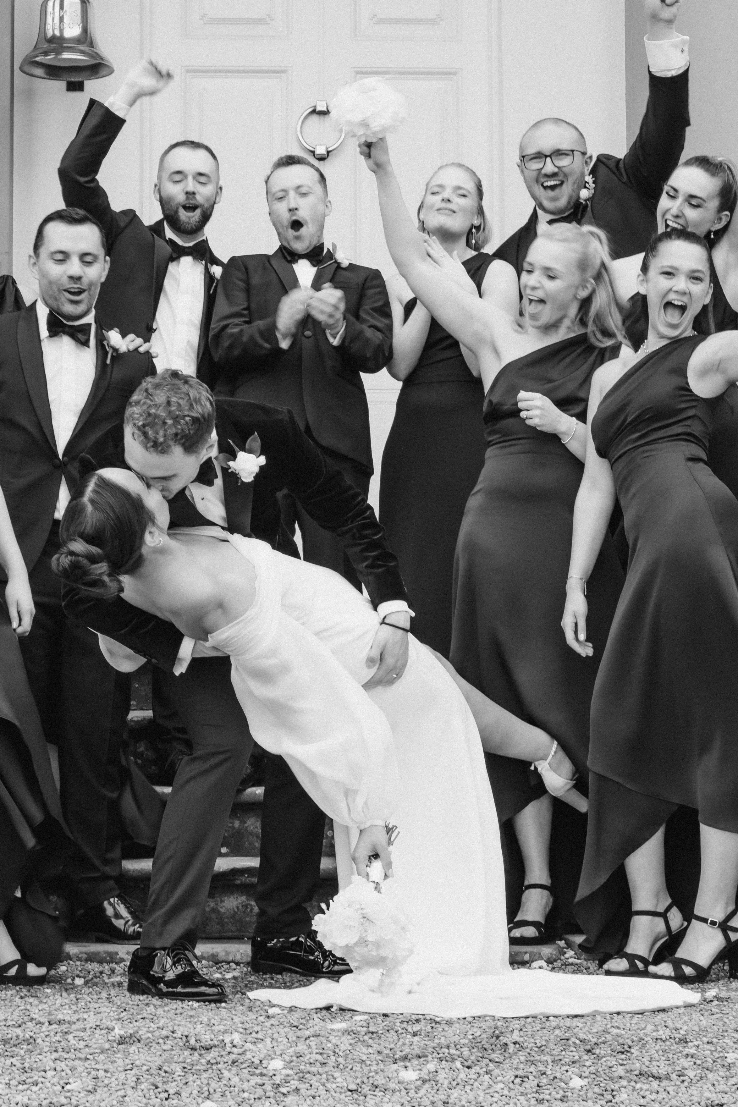 A black-and-white photo of a wedding celebration with a group of people celebrating outdoors, with a bride and groom kissing in front. The bride is holding a bouquet, and people around are smiling, cheering, and raising their hands in celebration.