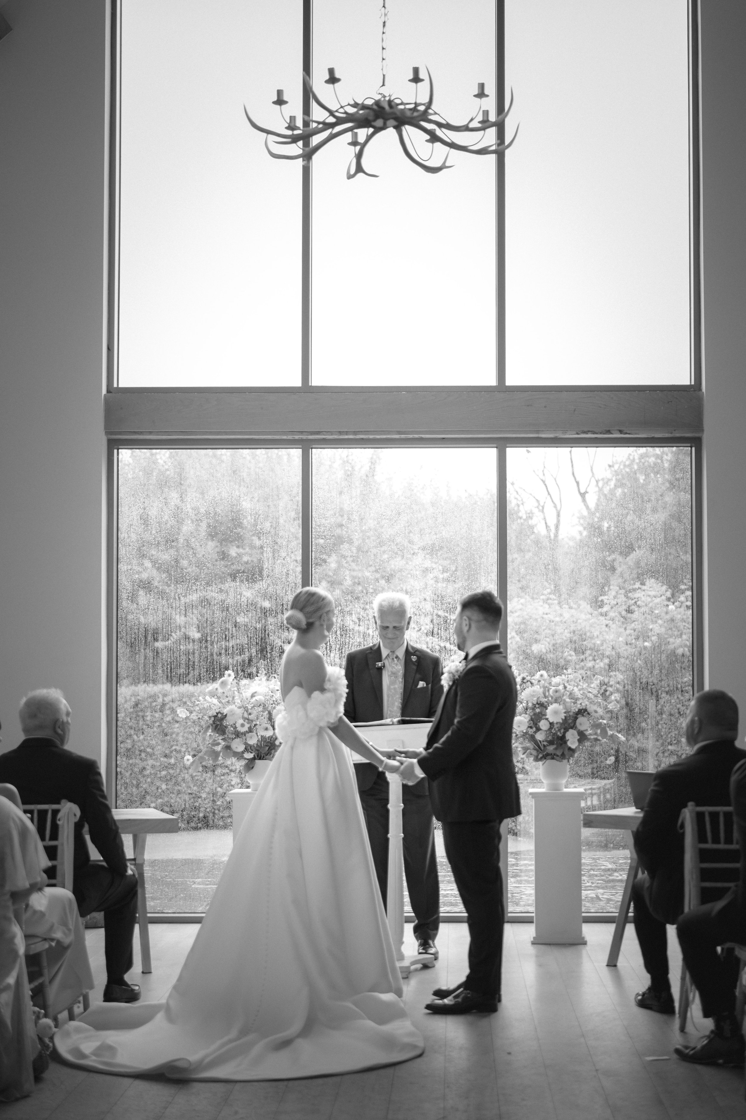 A wedding ceremony taking place indoors with a large window behind the couple and officiant. The bride and groom are holding hands, facing each other. The bride is in a long wedding gown and the groom in a dark suit. There are guests seated on either