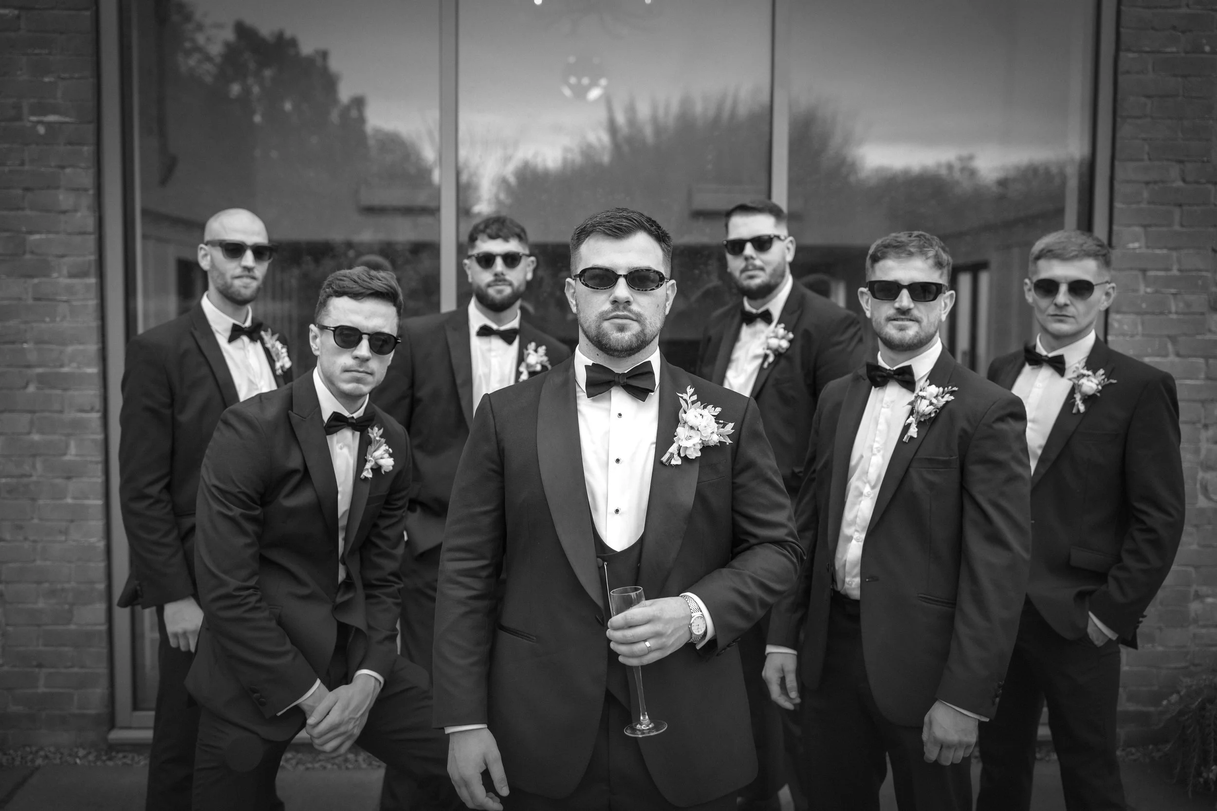 A group of seven men dressed in black tuxedos and sunglasses, posing outdoors in front of a building with brick and glass exterior, some with floral boutonnieres, one holding a champagne flute.