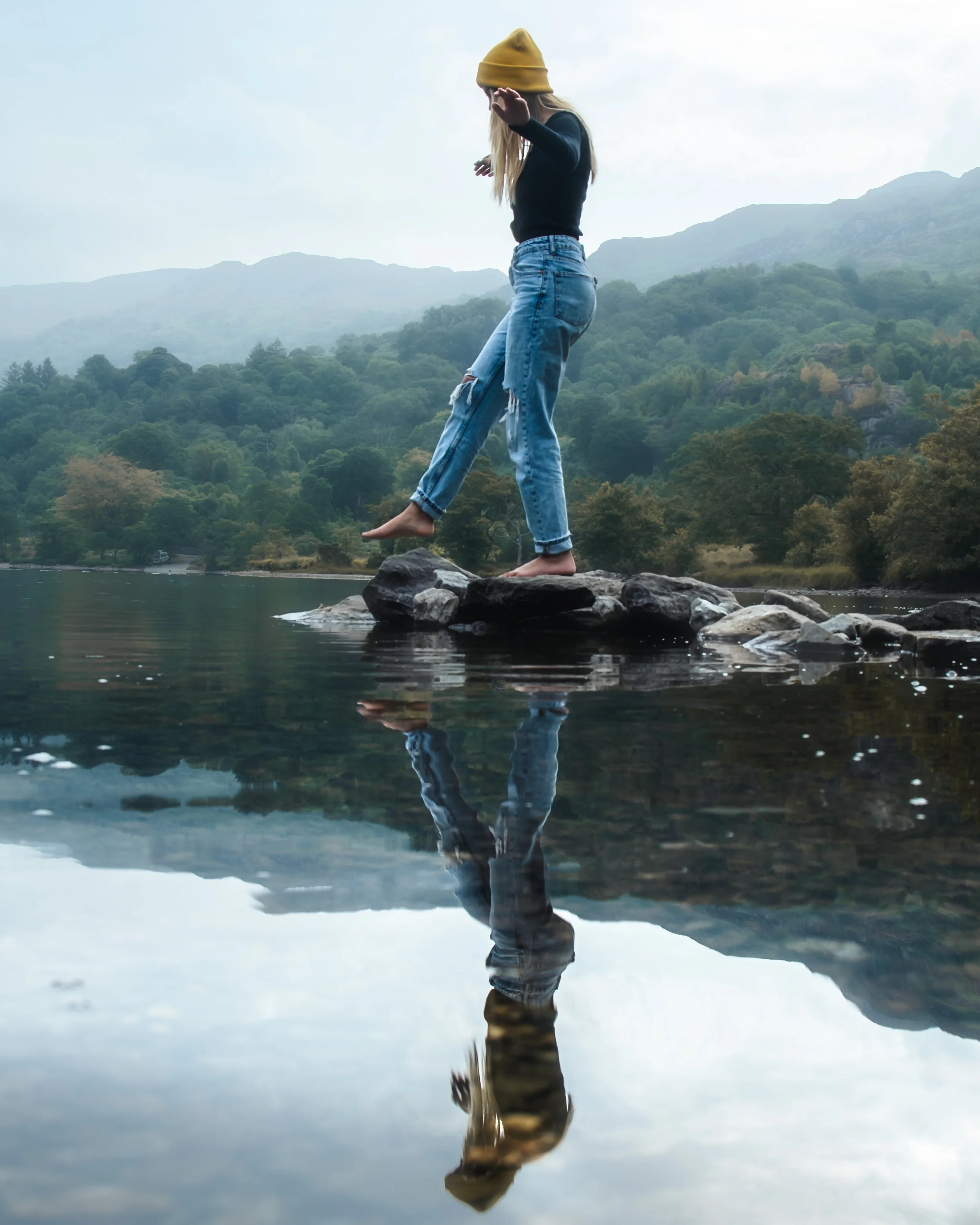 Young woman with a yellow beanie balancing on rocks in a river with mountains and trees in the background, and her reflection visible in the water.