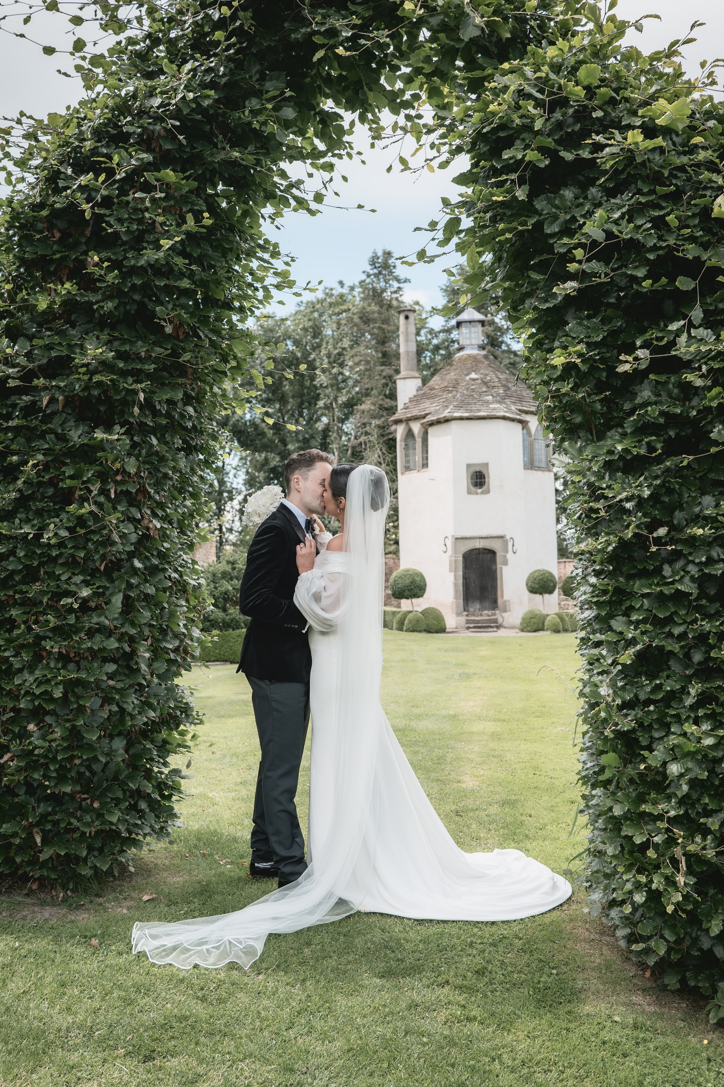 A bride and groom sharing a kiss under a leafy arch, with a small whimsical house in the background.