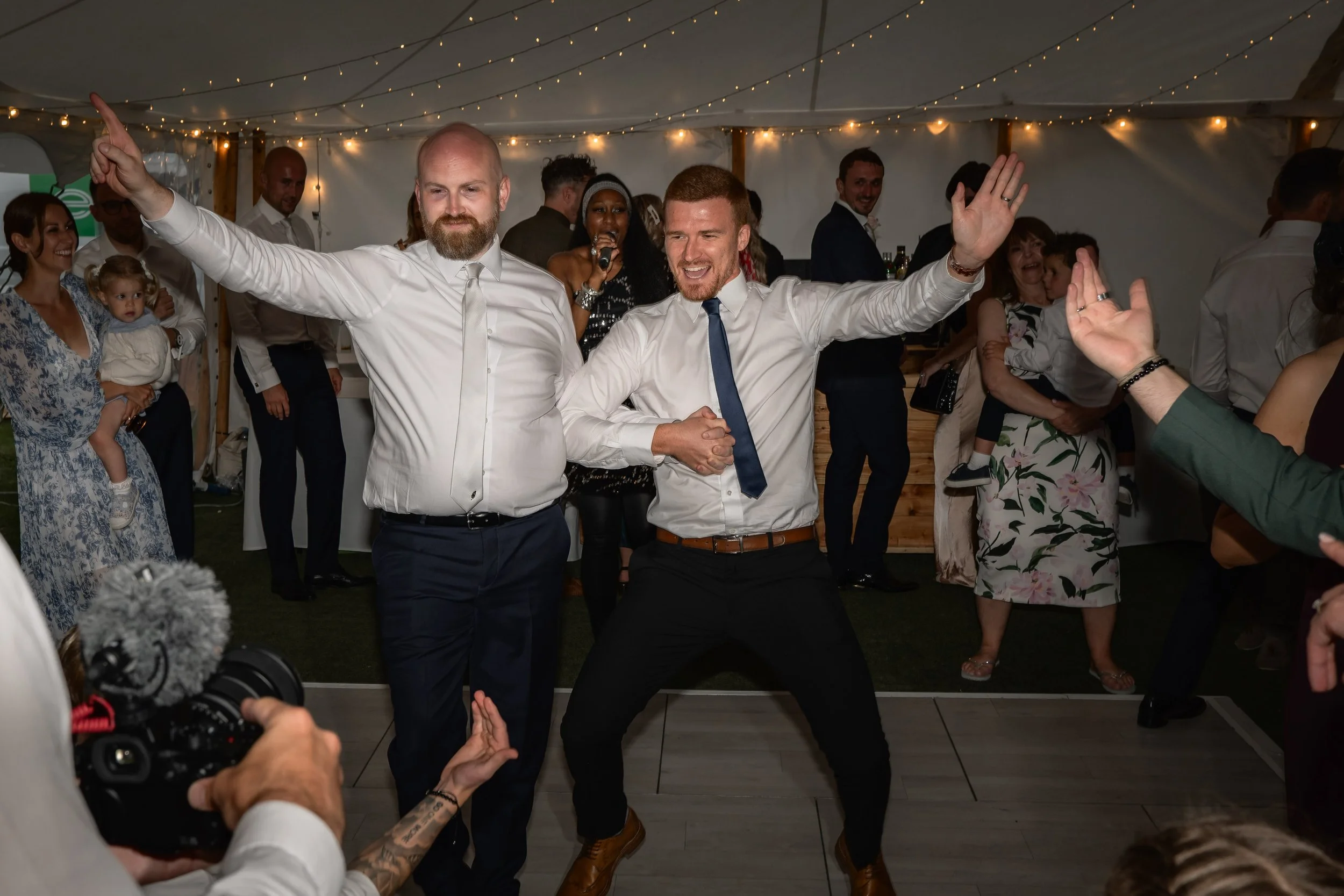 Two men dancing and holding hands at a celebration, surrounded by other guests under string lights.