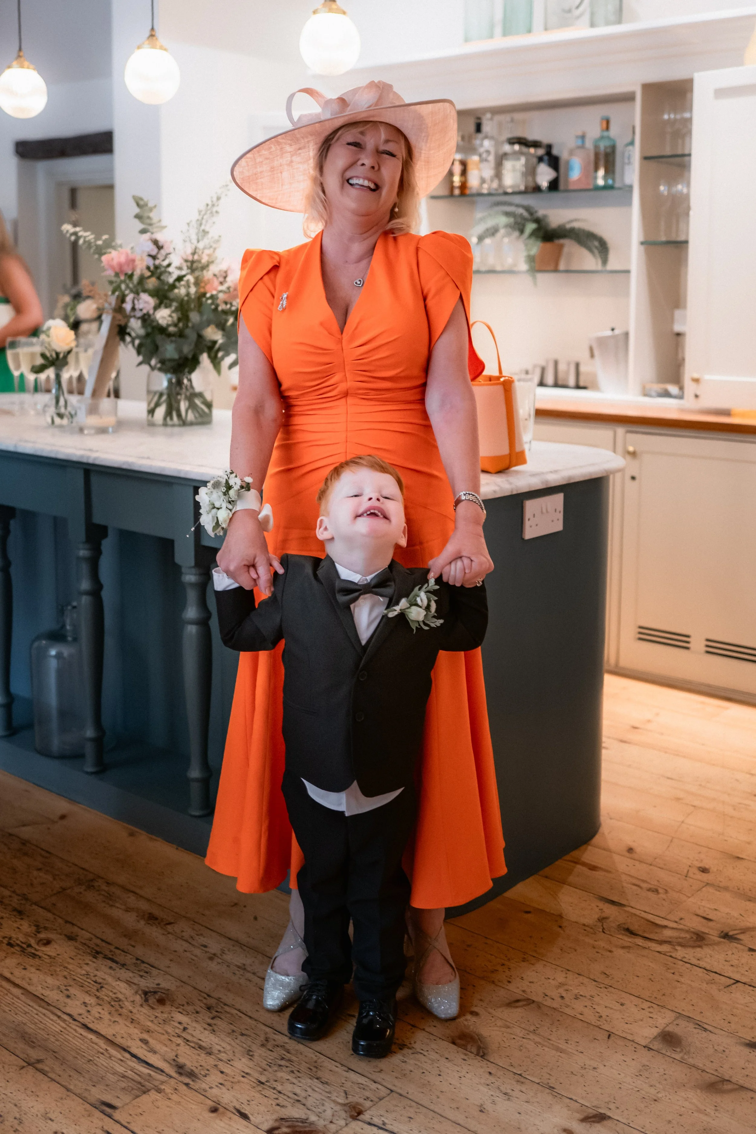 A woman in an orange dress and large hat is smiling while holding the hands of a young boy in a black tuxedo. They are standing indoors, with a decorated table and floral arrangements in the background, indicating a celebration or special event.