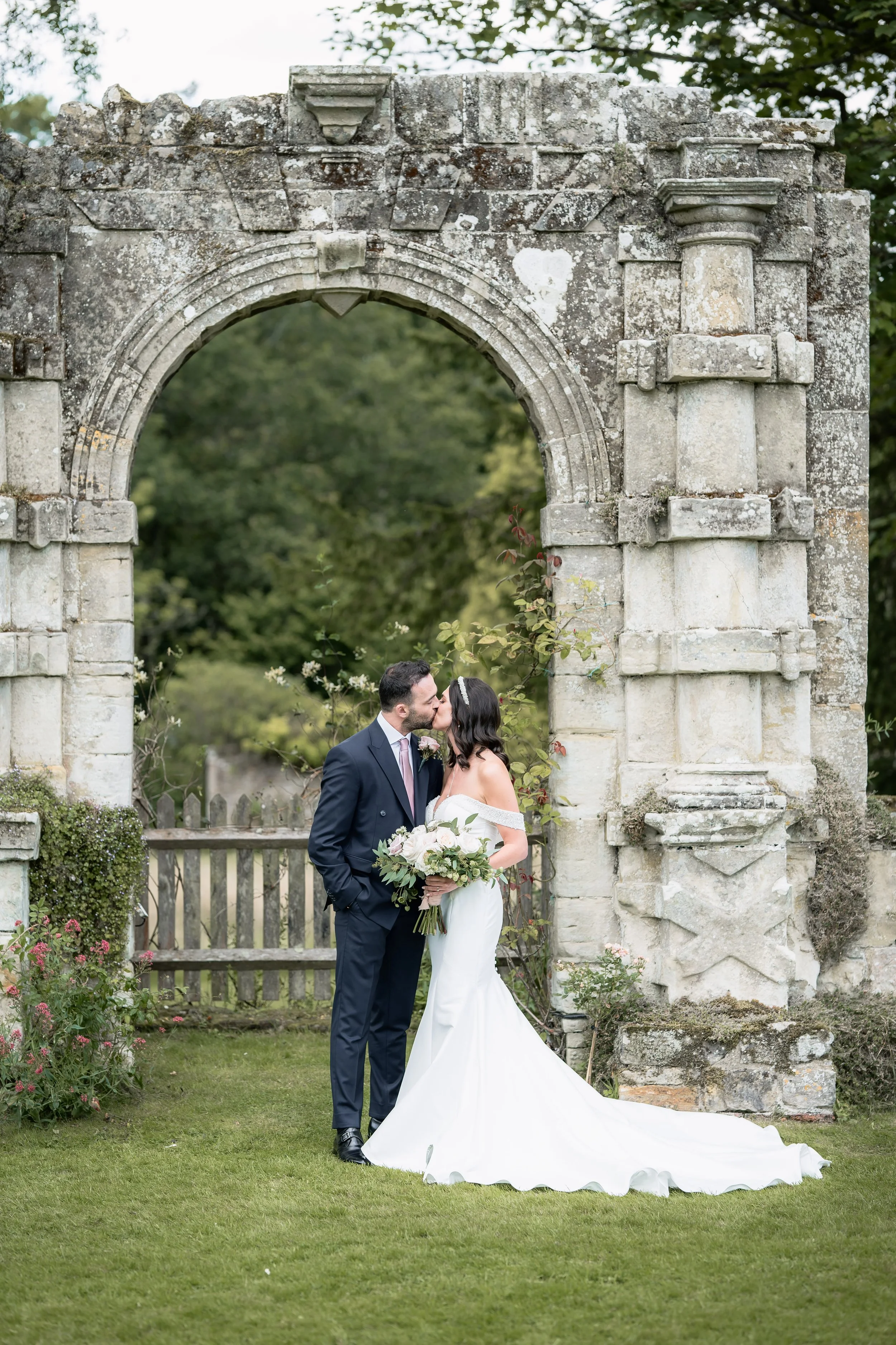 Bride and groom kissing under an old stone archway at their wedding, with greenery and flowers around.