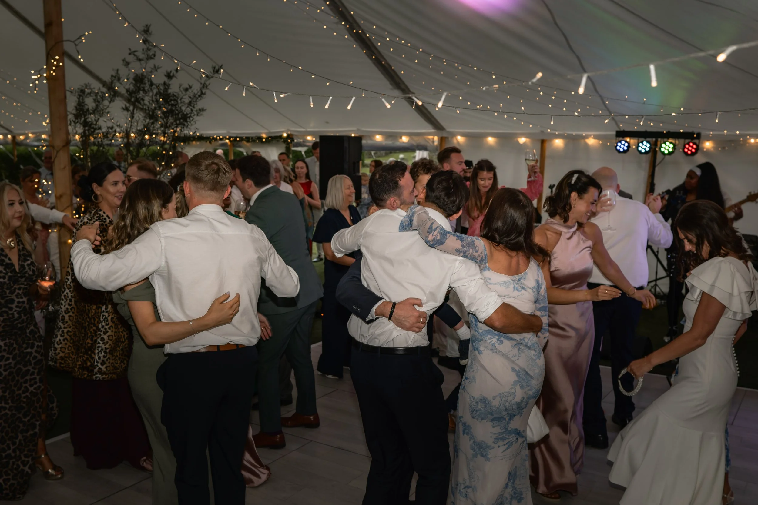 People dancing and celebrating at a wedding reception under a large tent decorated with string lights, with a band and musicians playing in the background.