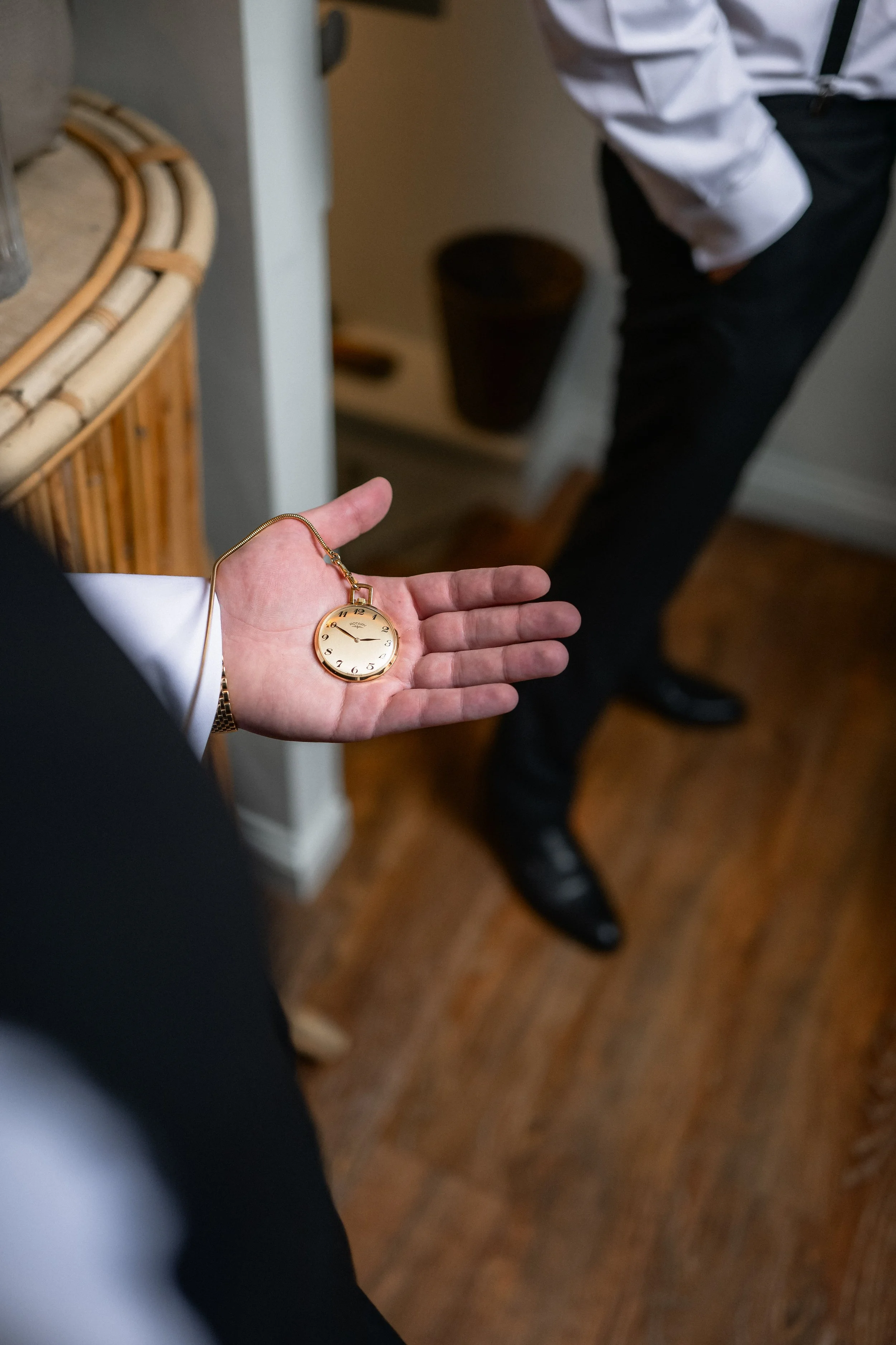 Person holding a pocket watch in their open palm, standing in a room with wooden flooring and another person dressed in formal attire standing nearby.