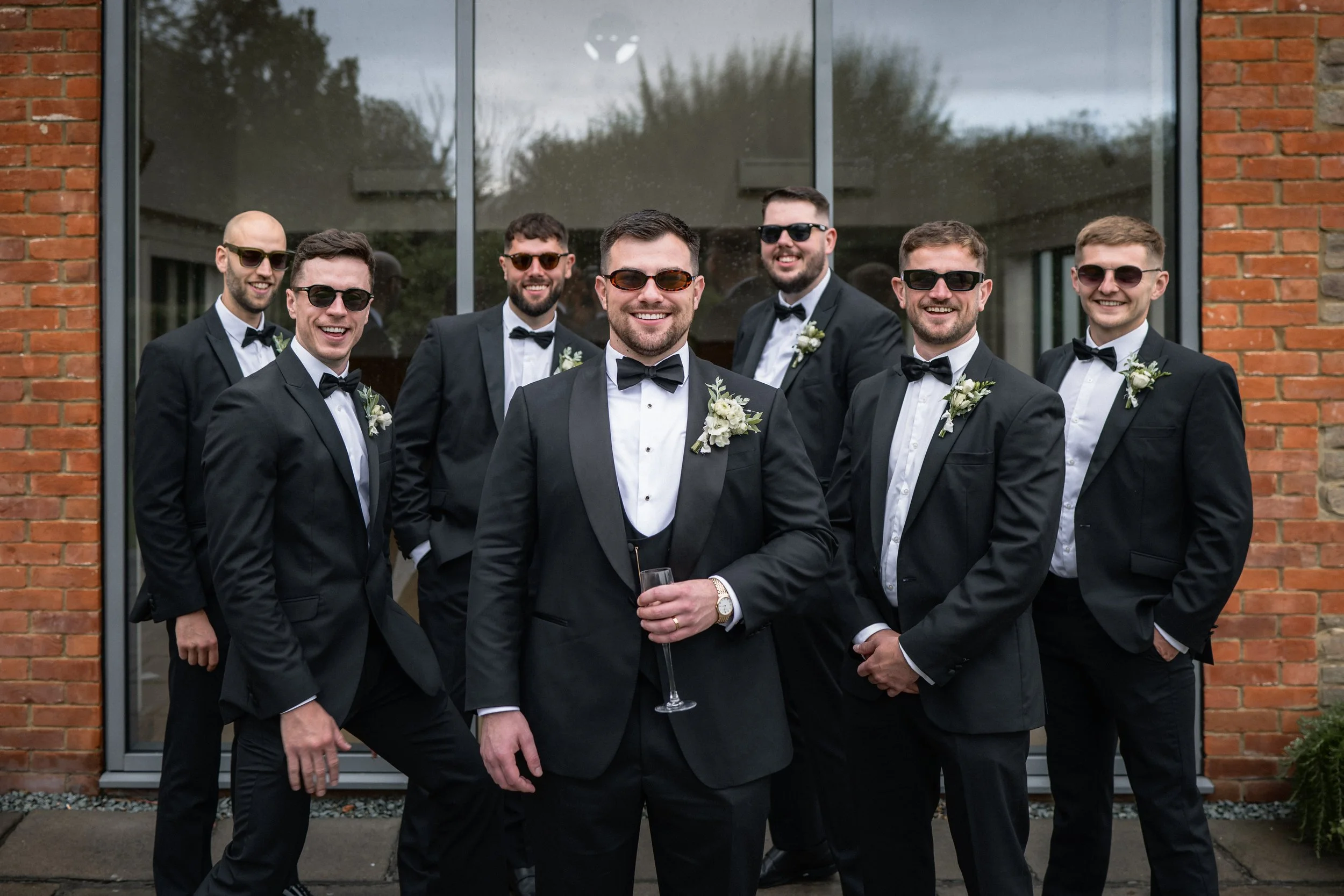 A group of seven men in black tuxedos, white shirts, and black bow ties posing in front of a glass window and brick wall. They are all wearing sunglasses and smiling, with some holding drinks. The man in the front center is holding a champagne flute 
