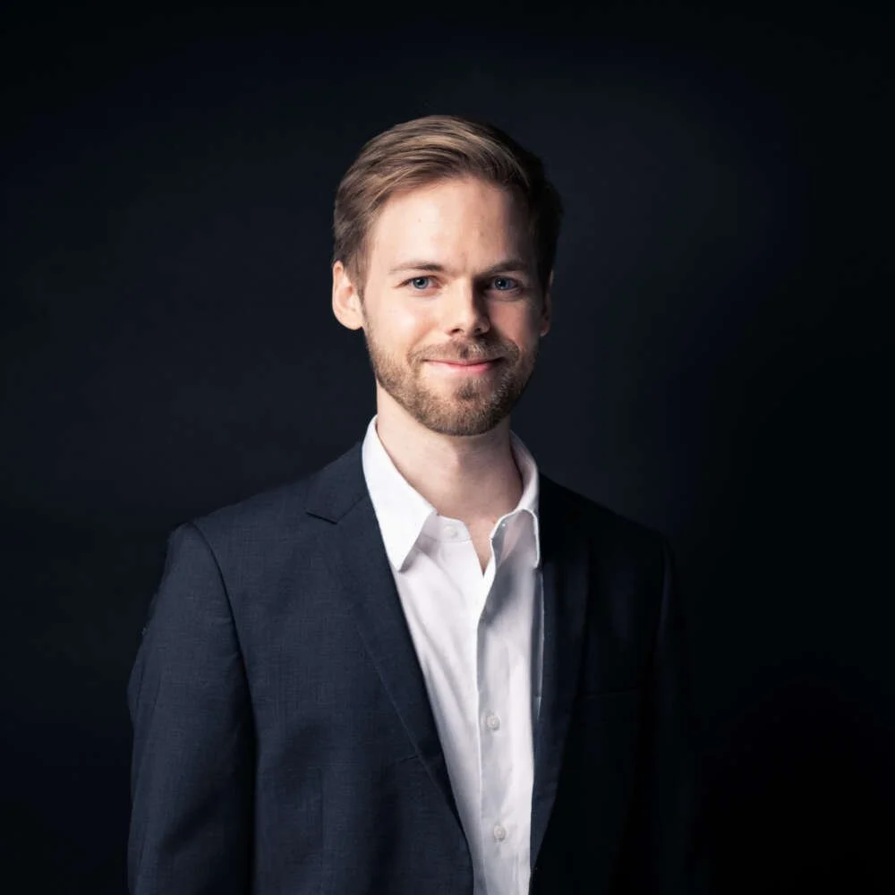 Portrait of a man with light brown hair and a beard, wearing a dark suit jacket and white shirt, against a dark background.