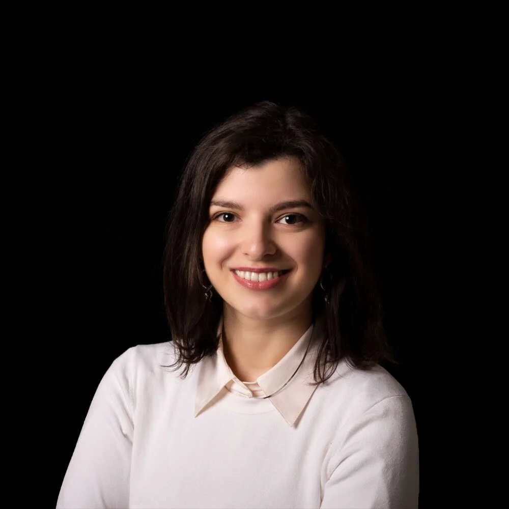 A young woman with dark hair, wearing a white shirt, smiling against a black background.
