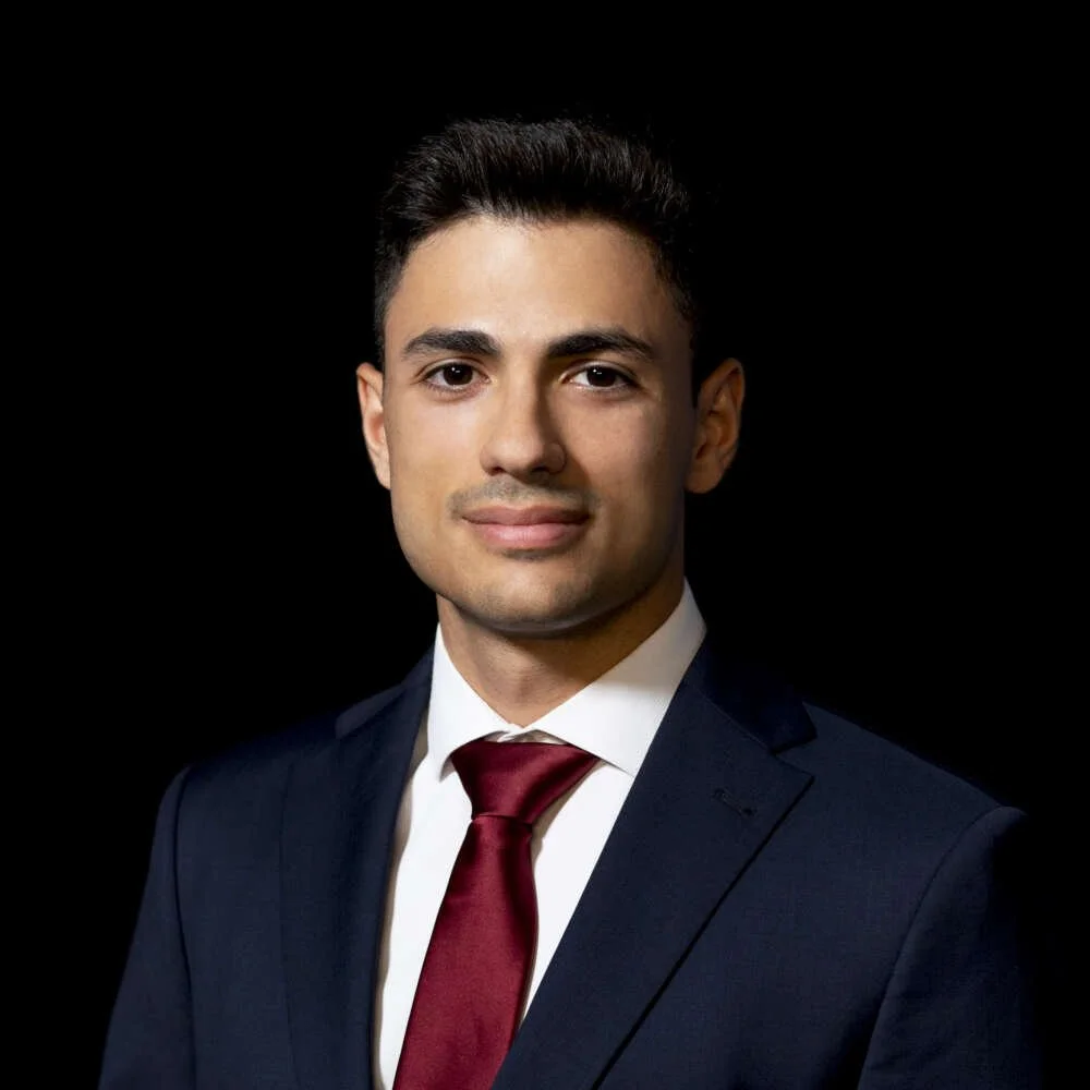 A portrait of a young man with dark hair, wearing a navy suit, white shirt, and red tie, against a black background.