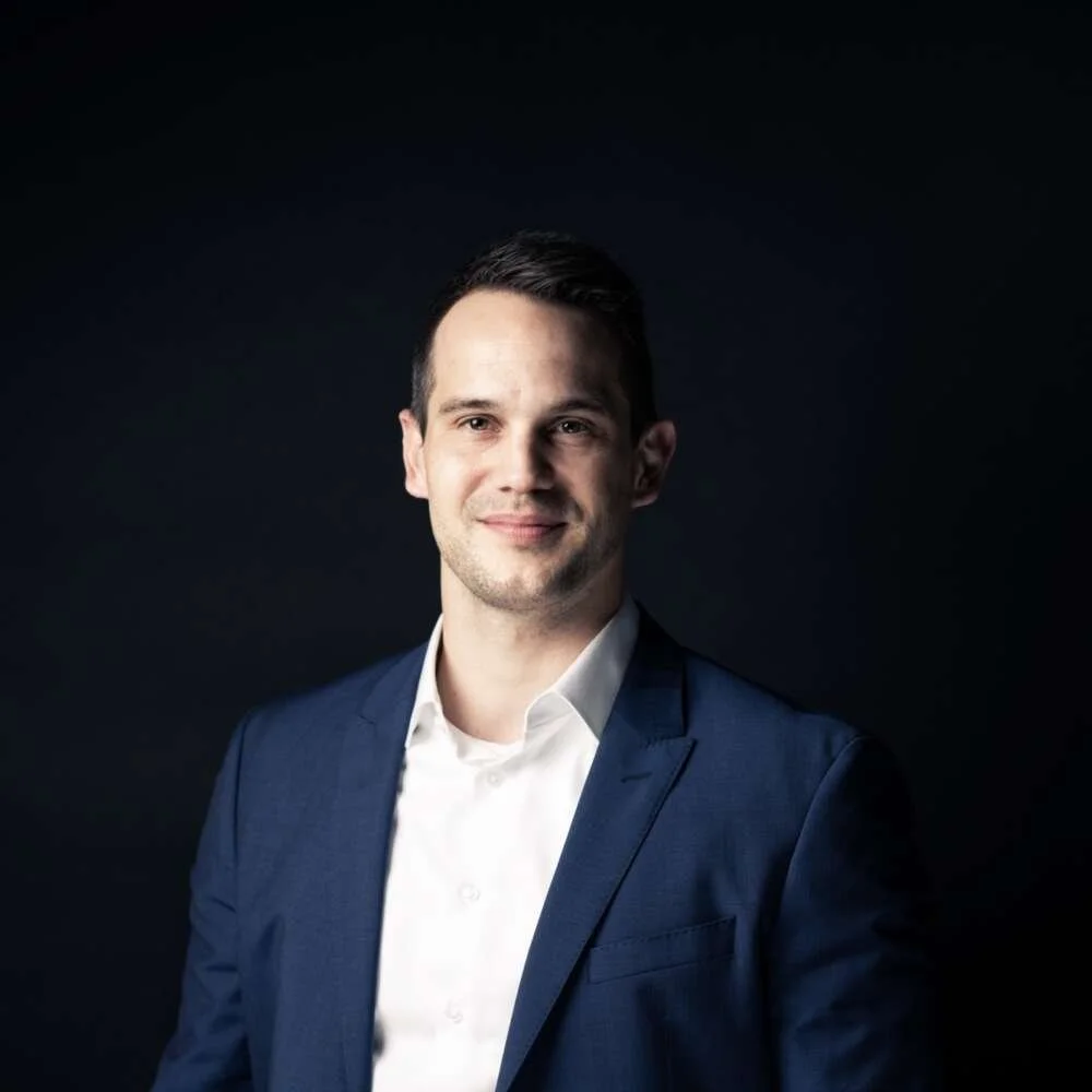 Portrait of a young man with dark hair wearing a navy suit and white shirt, smiling softly against a dark background.