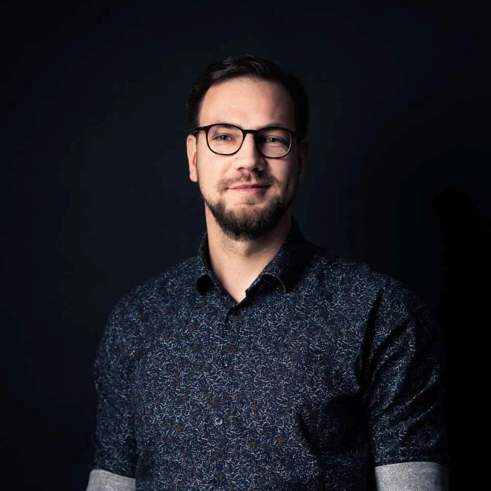 Portrait of a man with glasses and a beard, wearing a dark patterned shirt, against a dark background.