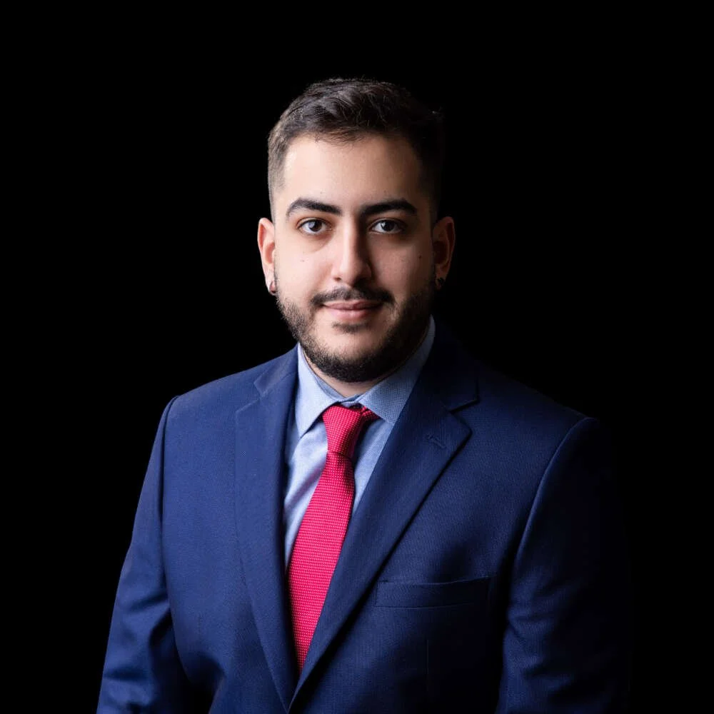 A portrait of a man with short dark hair and a beard, wearing a navy suit, light blue shirt, and red tie, against a black background.