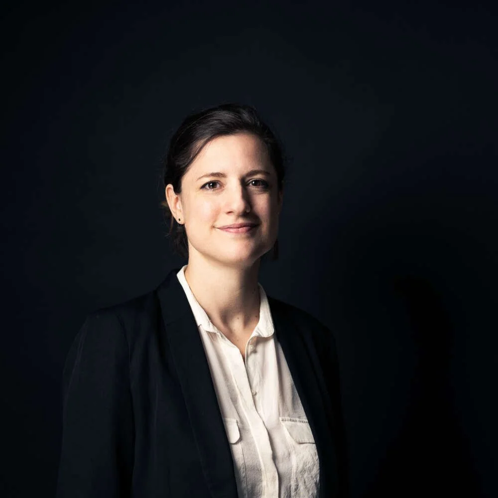 Headshot of a woman with short dark hair, wearing a dark blazer and a white blouse, standing against a dark background.