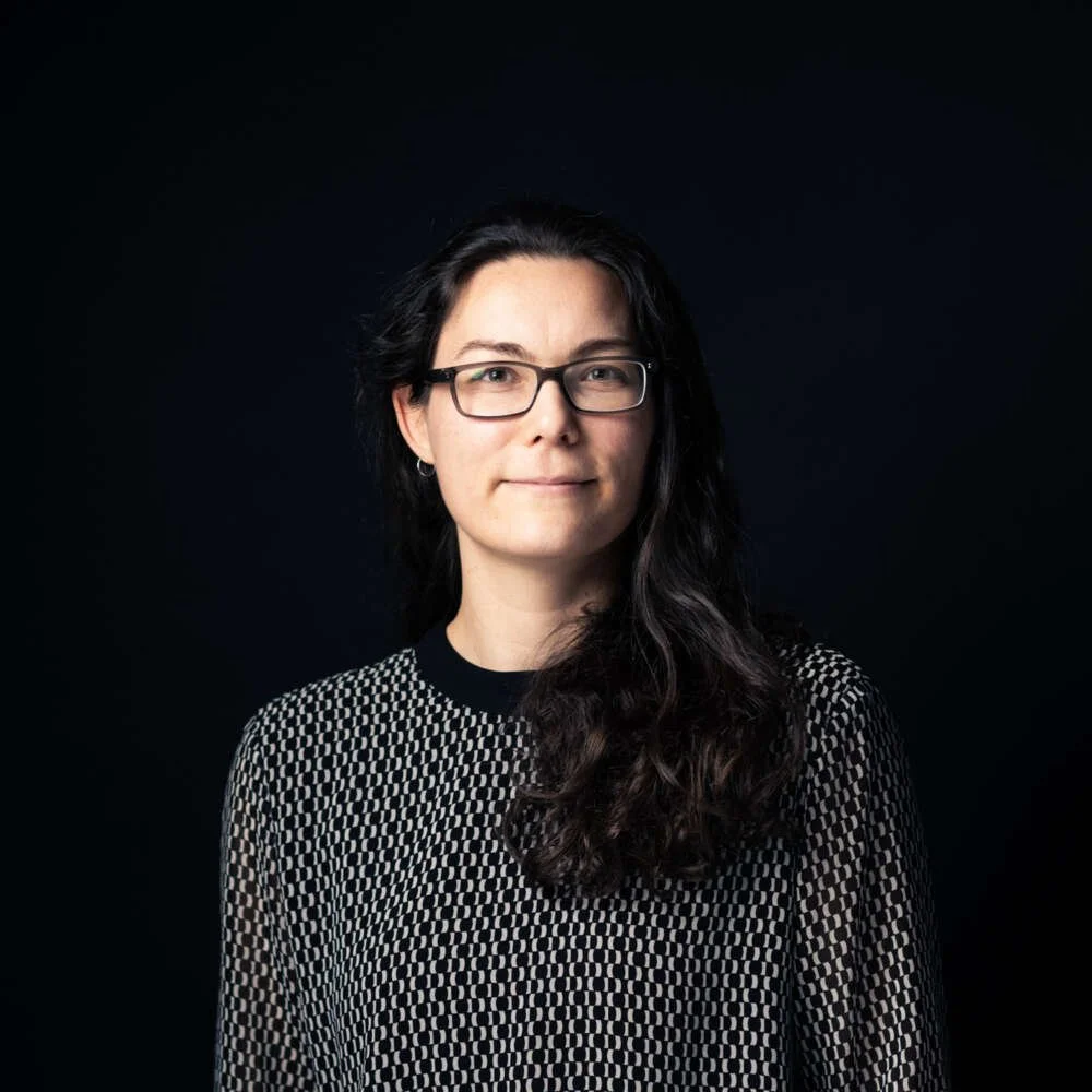 A woman with dark hair, glasses, and a patterned black and white top, standing against a black background.