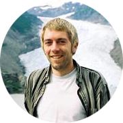 A smiling man with short blond hair and a beard, wearing a striped jacket over a white shirt, standing in front of a waterfall.