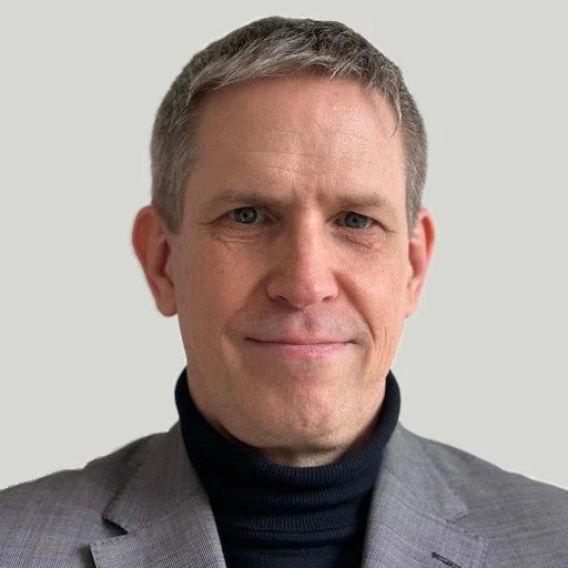 A professional man with short gray hair, blue eyes, wearing a light gray suit jacket and a black turtleneck, smiling slightly against a plain light background.