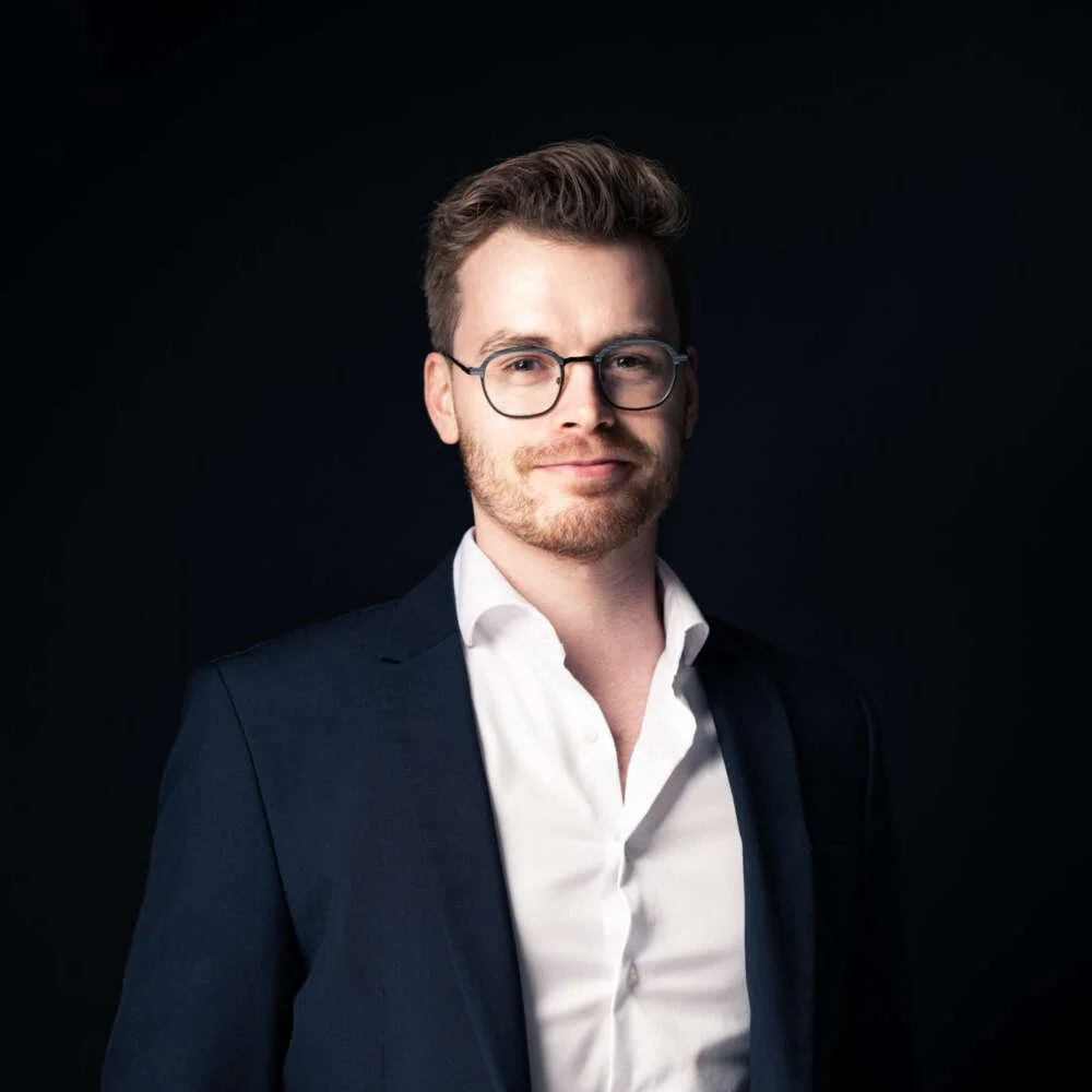 A man with glasses, a beard, and brown hair, wearing a dark blazer and white shirt, standing against a black background.