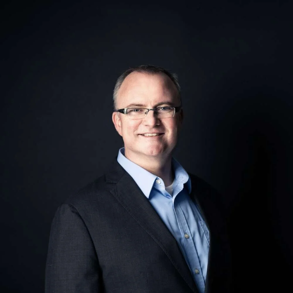A professional headshot of a smiling man wearing glasses, a blue collared shirt, and a dark blazer against a dark background.
