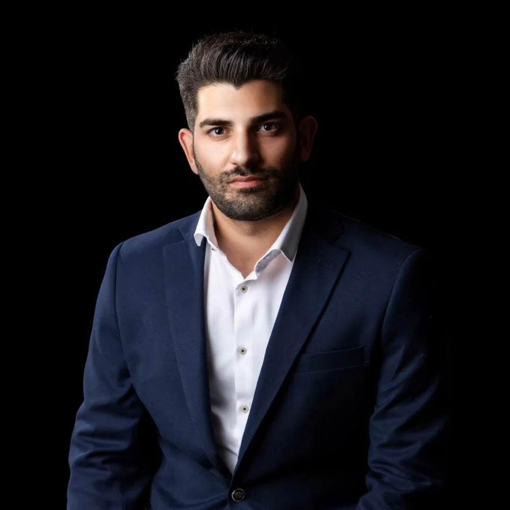 A man with dark hair, a beard, and wearing a dark suit and white shirt, standing against a black background.