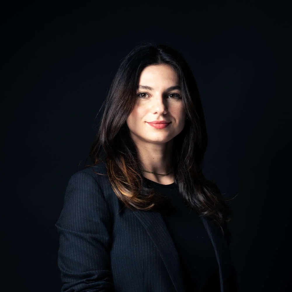 Portrait of a woman with dark hair wearing a dark blazer against a black background.