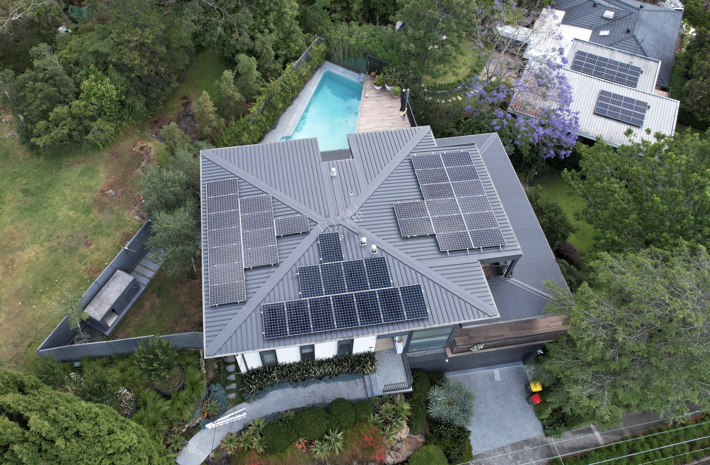 An aerial view of a house featuring a grey metal roof with multiple solar panels, a backyard with a swimming pool, surrounded by lush green trees and plants.